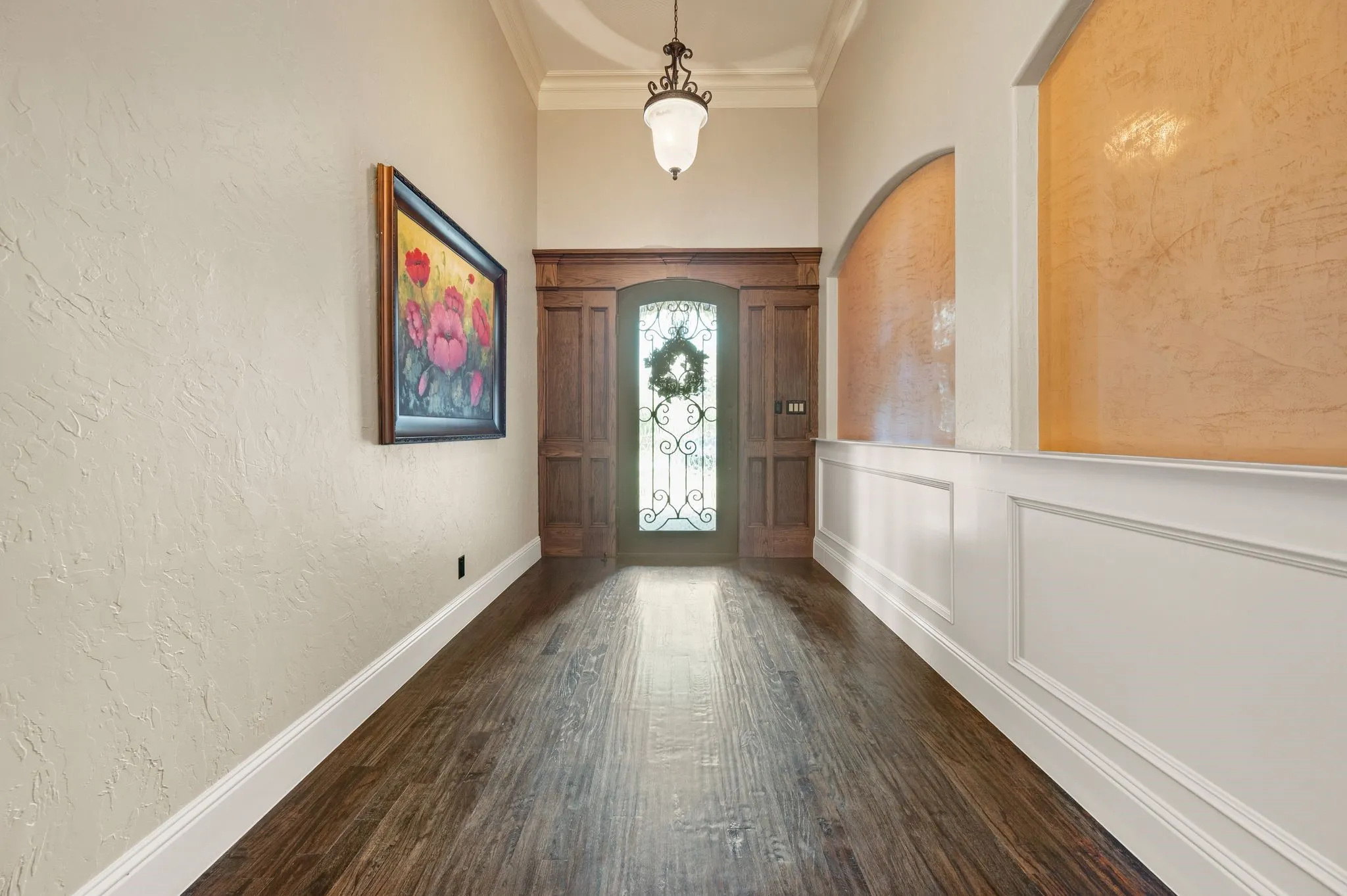 Wood-finish flooring, wainscoting, arched wall recesses with textured finish, decorative entry door with wrought iron detailing, and an overhead pendant light fixture