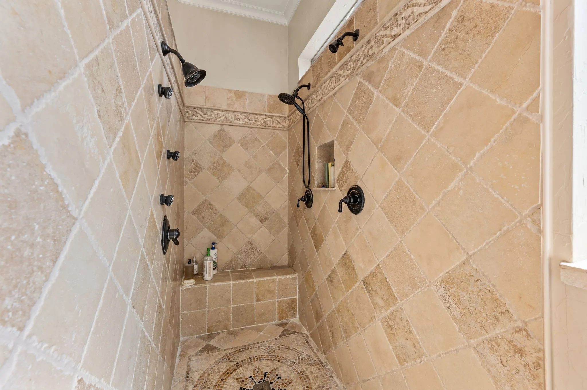 Expansive walk-in shower featuring multiple showerheads, a built-in bench, decorative tile accents, and an integrated niche