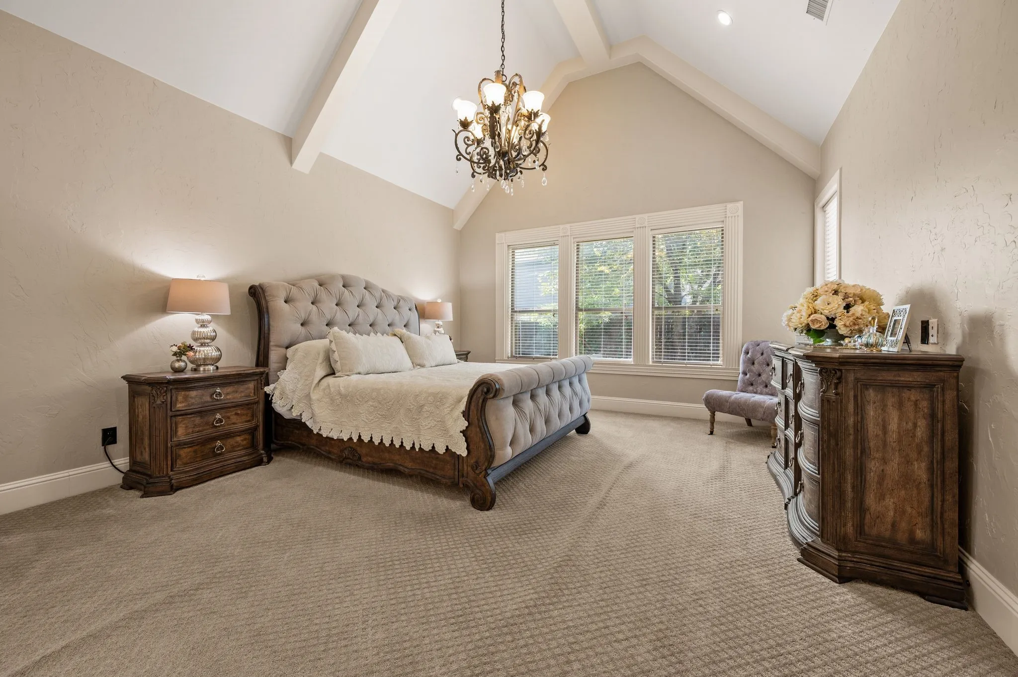 Vaulted ceiling with exposed beams, decorative chandelier, large windows with blinds, and neutral-toned wall finish