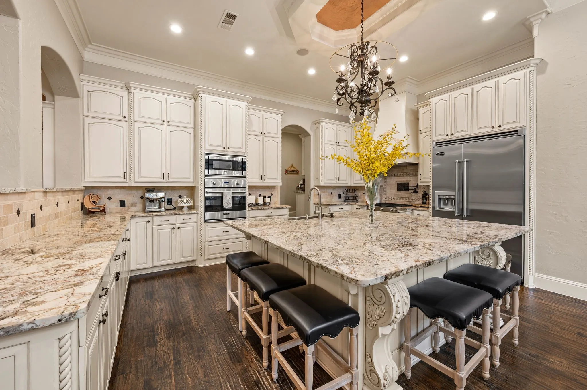 Gourmet kitchen featuring white cabinetry, stainless steel appliances, a large island with seating, dark wood-finish flooring, and a coffered ceiling with a chandelier
