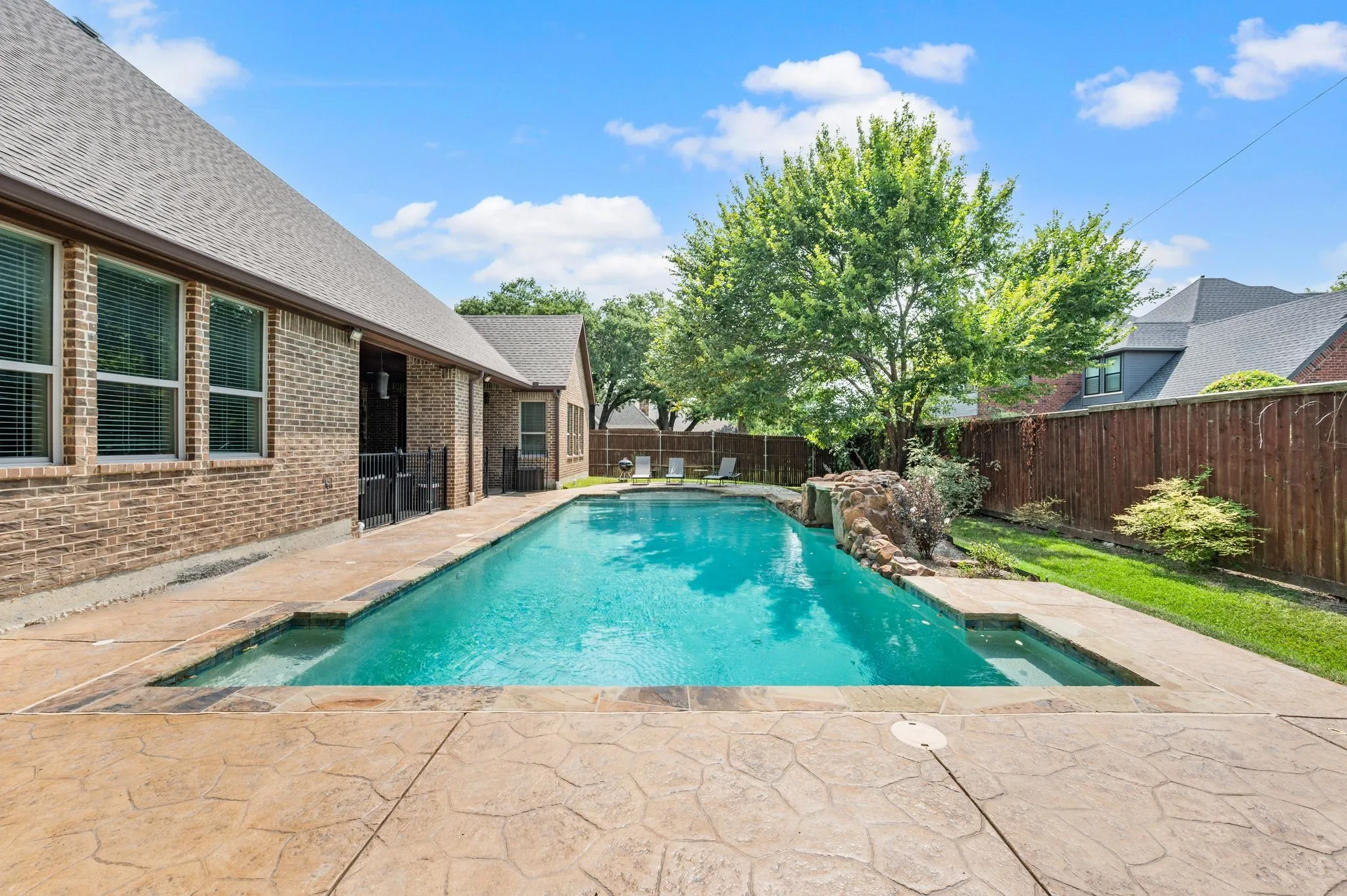 Rectangular swimming pool with a stone waterfall feature, surrounded by stamped concrete decking and a brick exterior