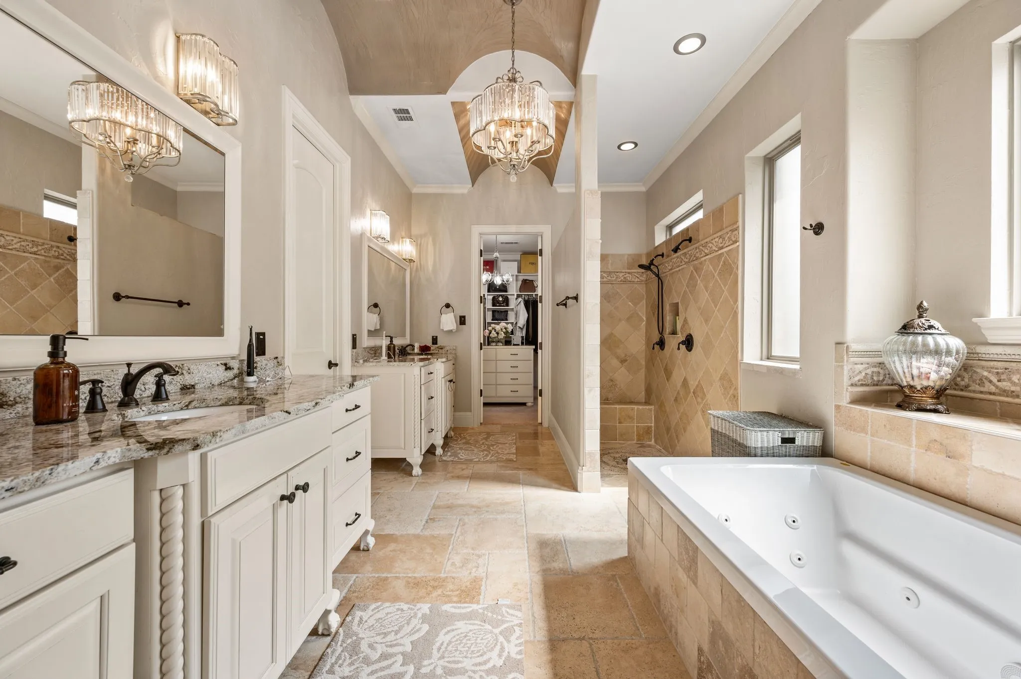 Dual vanities with granite countertops, jetted soaking tub, walk-in shower with tile surround, and vaulted ceiling with chandelier