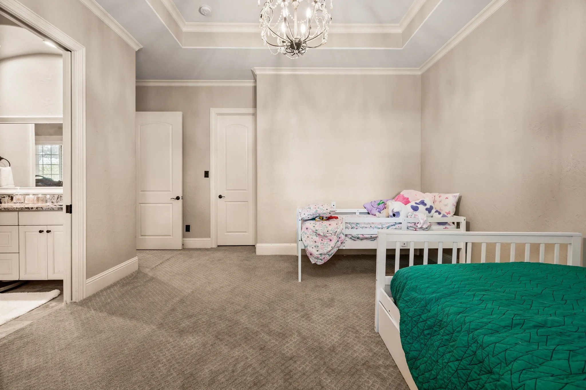 Spacious room featuring tray ceiling, chandelier, soft carpeting, and an adjoining bath with vanity and stone-finish countertop