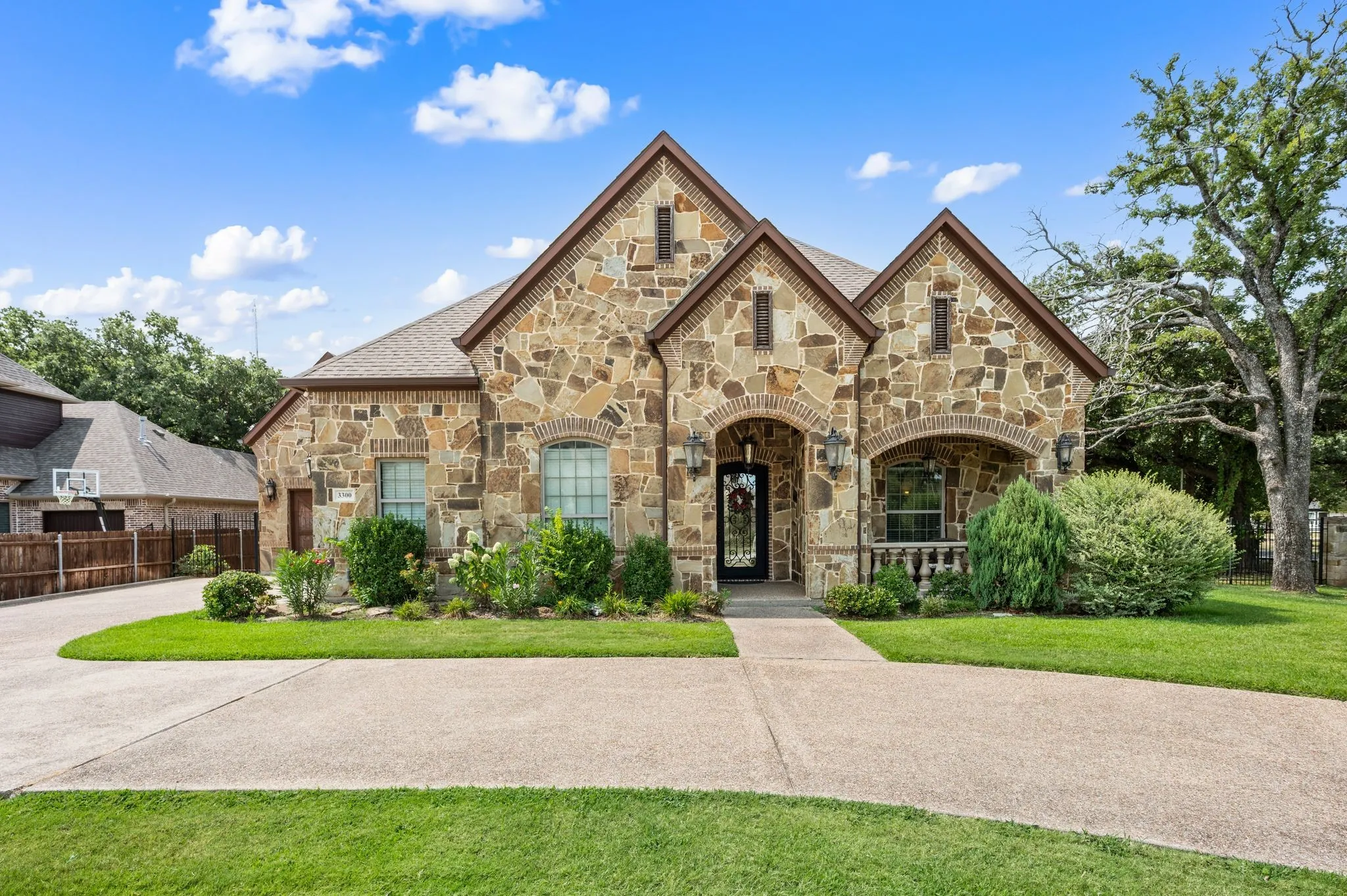 Stone facade with arched entries, a gabled roofline, mature landscaping, and a concrete driveway
