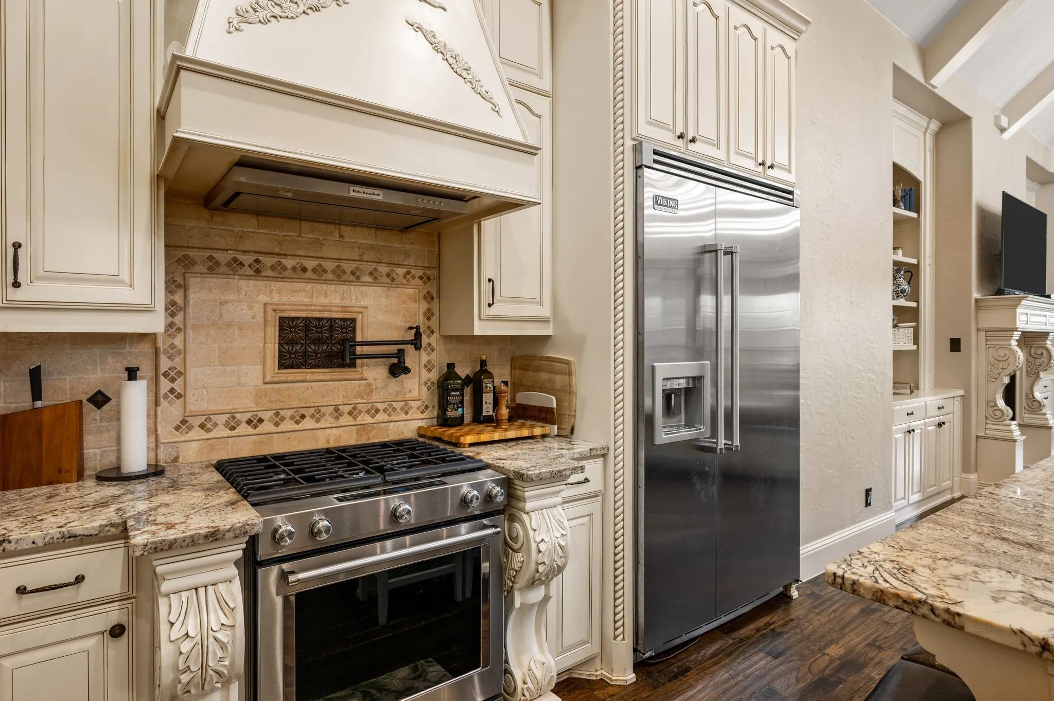 Stainless steel gas range with a decorative tile backsplash, custom range hood, built-in refrigerator, and stone countertops