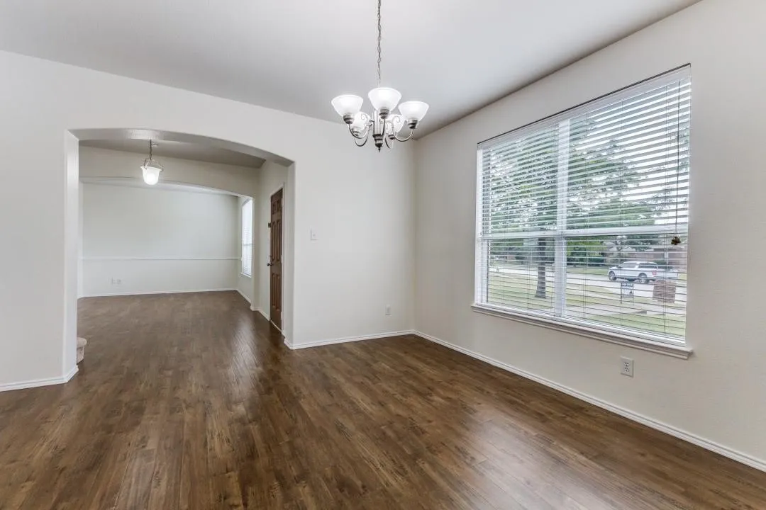 Spacious room with wood-finish flooring, a large window with blinds, and a five-light chandelier