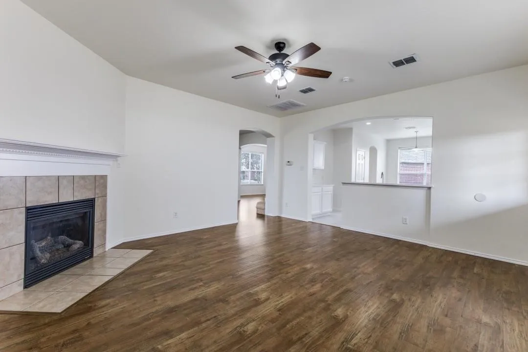 Wood-finish flooring throughout, a tiled fireplace with mantel, ceiling fan with integrated lighting, and arched pass-through to a separate area featuring white cabinetry