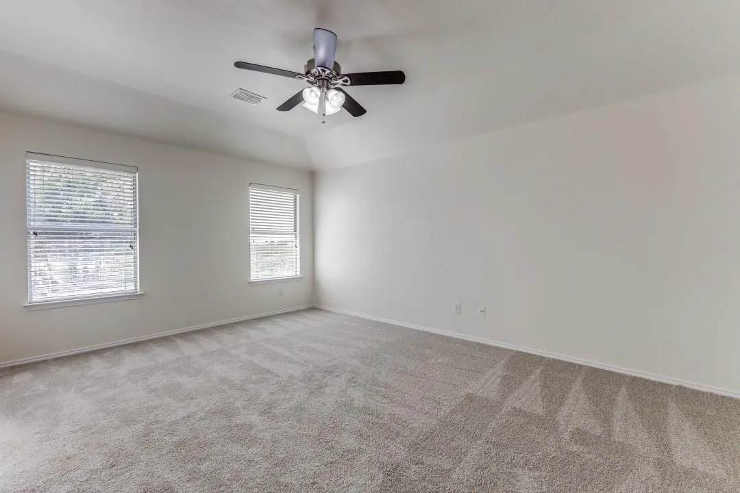 Spacious room featuring neutral carpeting, two windows with blinds, a ceiling fan with integrated lighting, and a tray ceiling