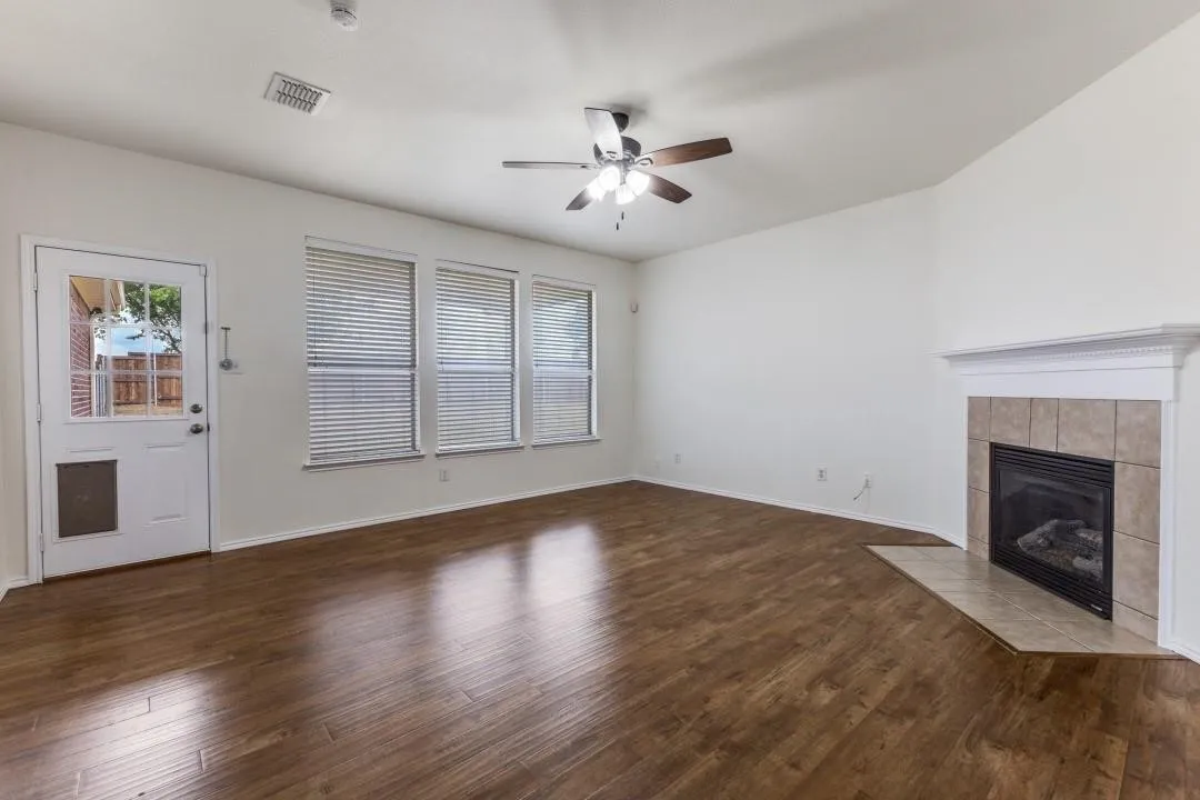 Wood-finish flooring throughout, a gas fireplace with tile surround, multiple windows with blinds, a ceiling fan, and a glass-paneled exterior door