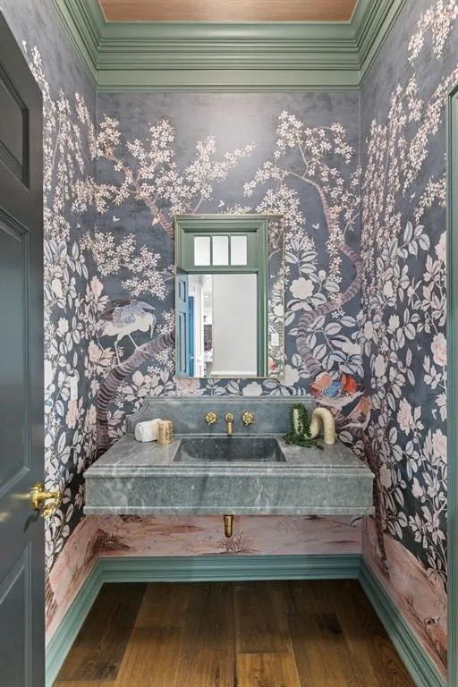 Wood-finish flooring, ornate wall covering, vanity with integrated basin, polished brass fixtures, and a mirrored cabinet
