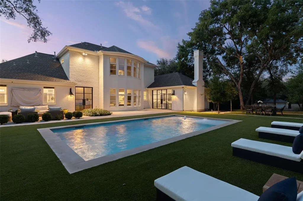 White brick exterior, rectangular swimming pool, expansive artificial turf lawn, mature trees, and a tall exterior chimney