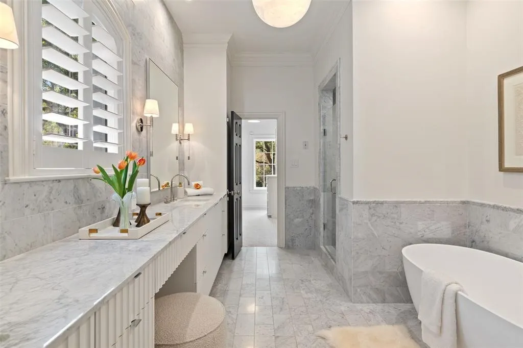 Window with plantation shutters, double vanity with dual sinks, built-in bathtub, glass-enclosed shower, and stone tile flooring