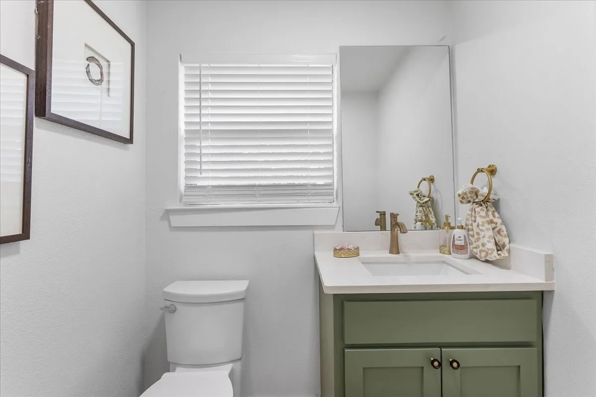 Half Bathroom near kitchen with white toilet, vanity with sage green cabinetry, integrated sink, and bronze-finish faucet, accompanied by a wall-mounted mirror