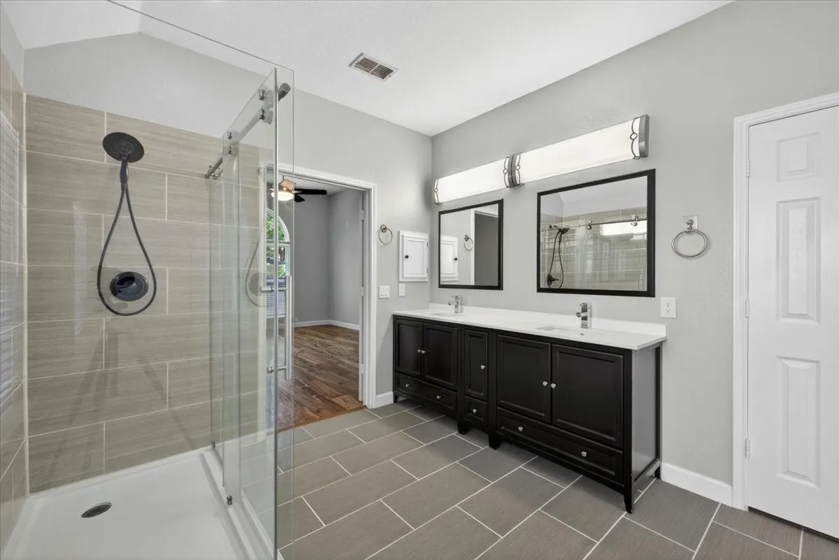 Spa-inspired primary bathroom featuring a frameless glass-enclosed walk-in shower with herringbone tile surround, dual vanities with black cabinetry and white quartz countertops, polished chrome fixtures, and tile flooring throughout