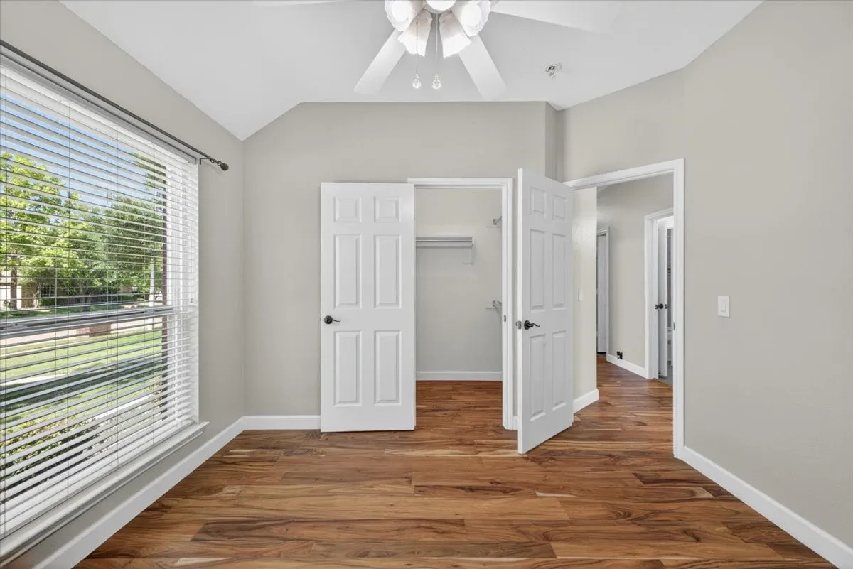Wood-finish flooring, vaulted ceiling, white ceiling fan, white paneled doors, and a large window with blinds