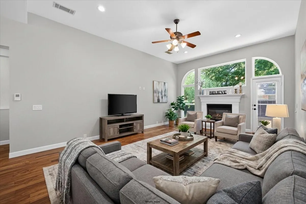 Vaulted ceiling with a ceiling fan, hardwood flooring, fireplace with a white mantel, arched and rectangular windows, and recessed lighting