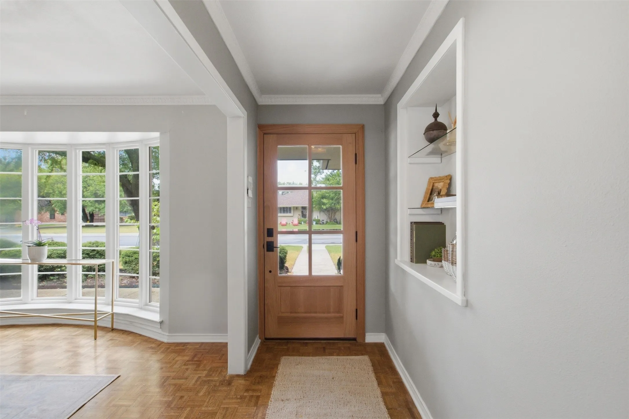 Wood-finish parquet flooring, bay window with multiple panes, natural wood entry door with glass inserts, recessed built-in shelving, and crown molding details