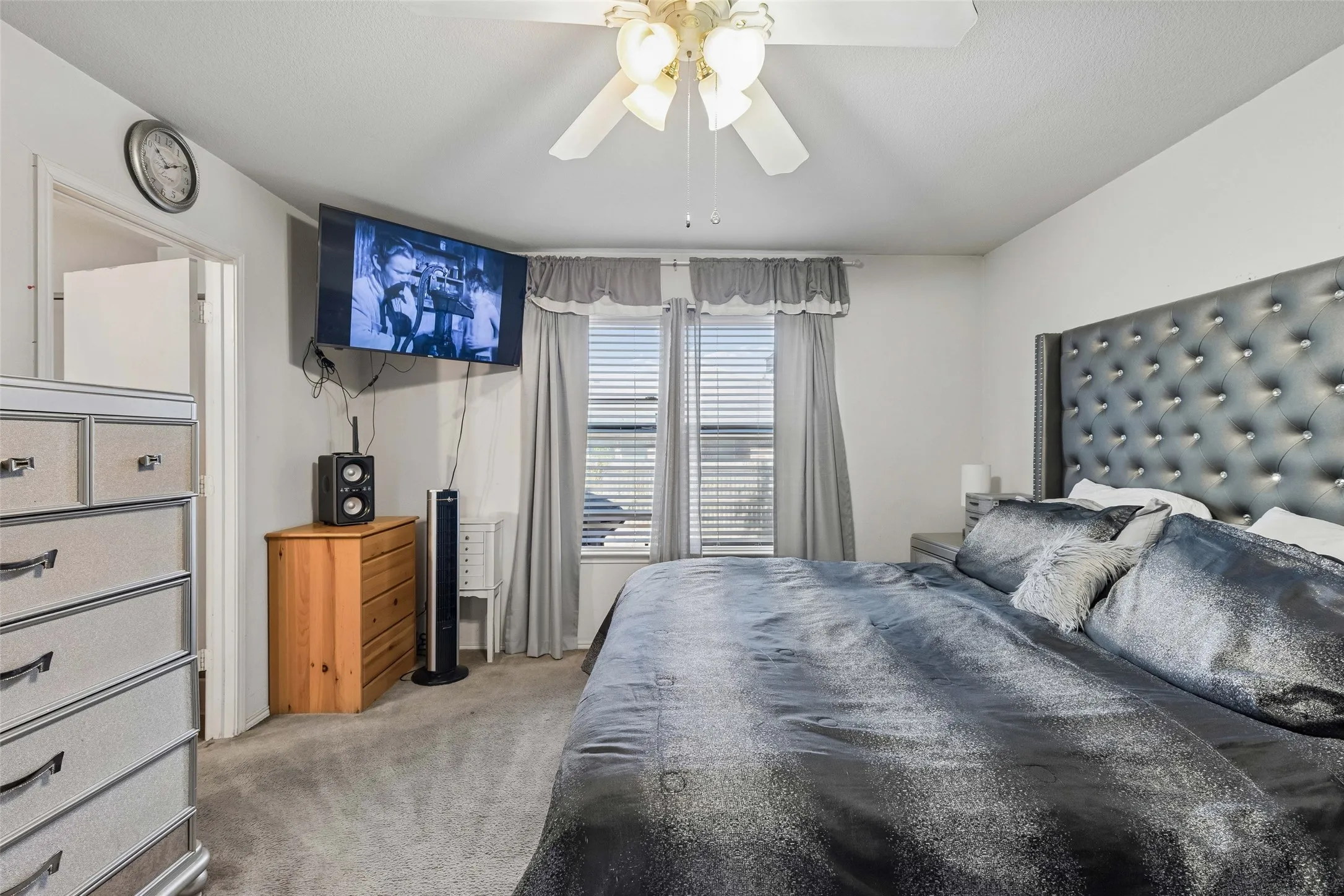 Carpeted room with a ceiling fan, two windows featuring blinds and curtains, and a tufted headboard