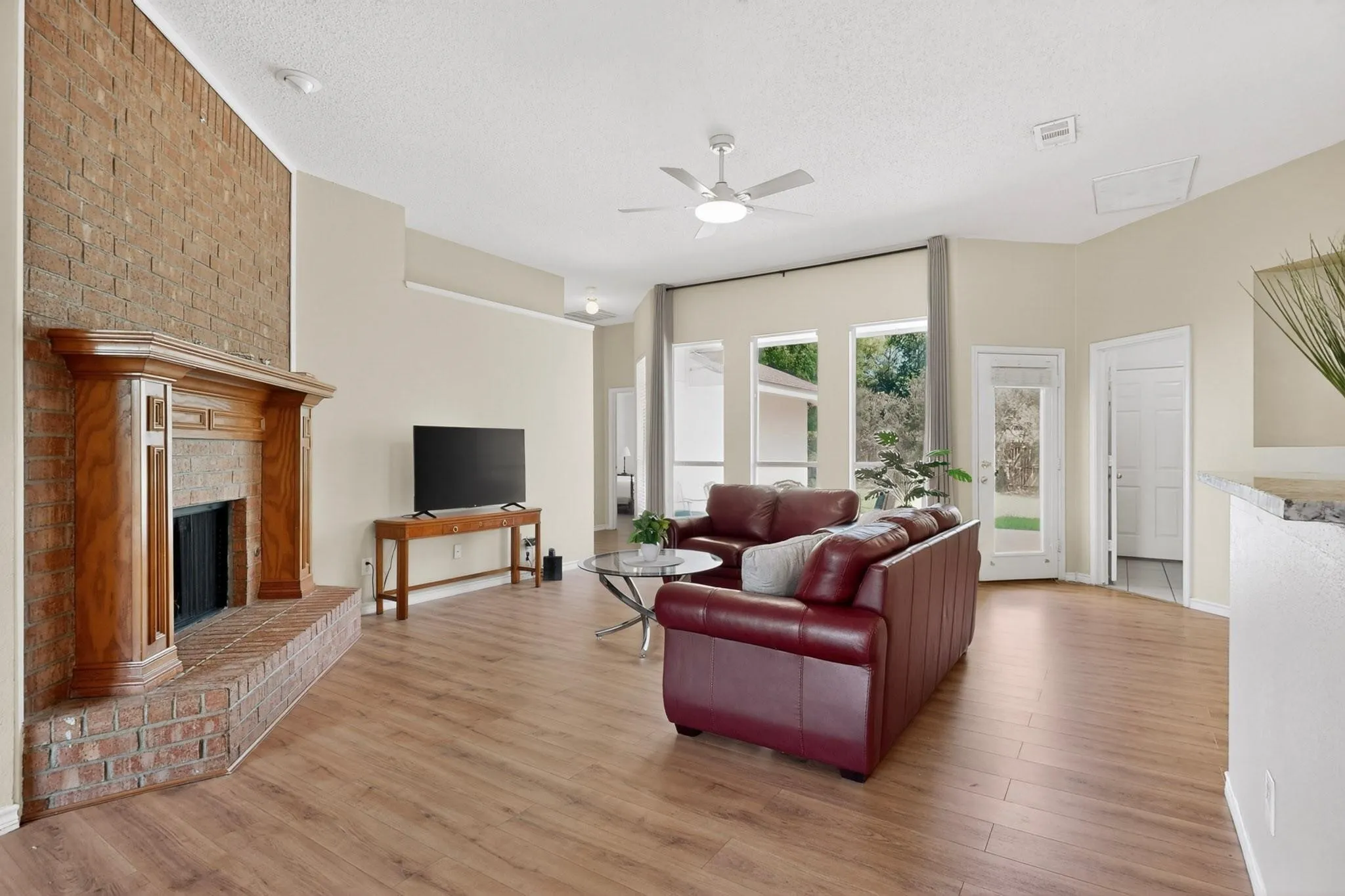 Living area featuring wood-finish flooring, a brick-surround fireplace with a wood mantle, a ceiling fan, and multiple large windows