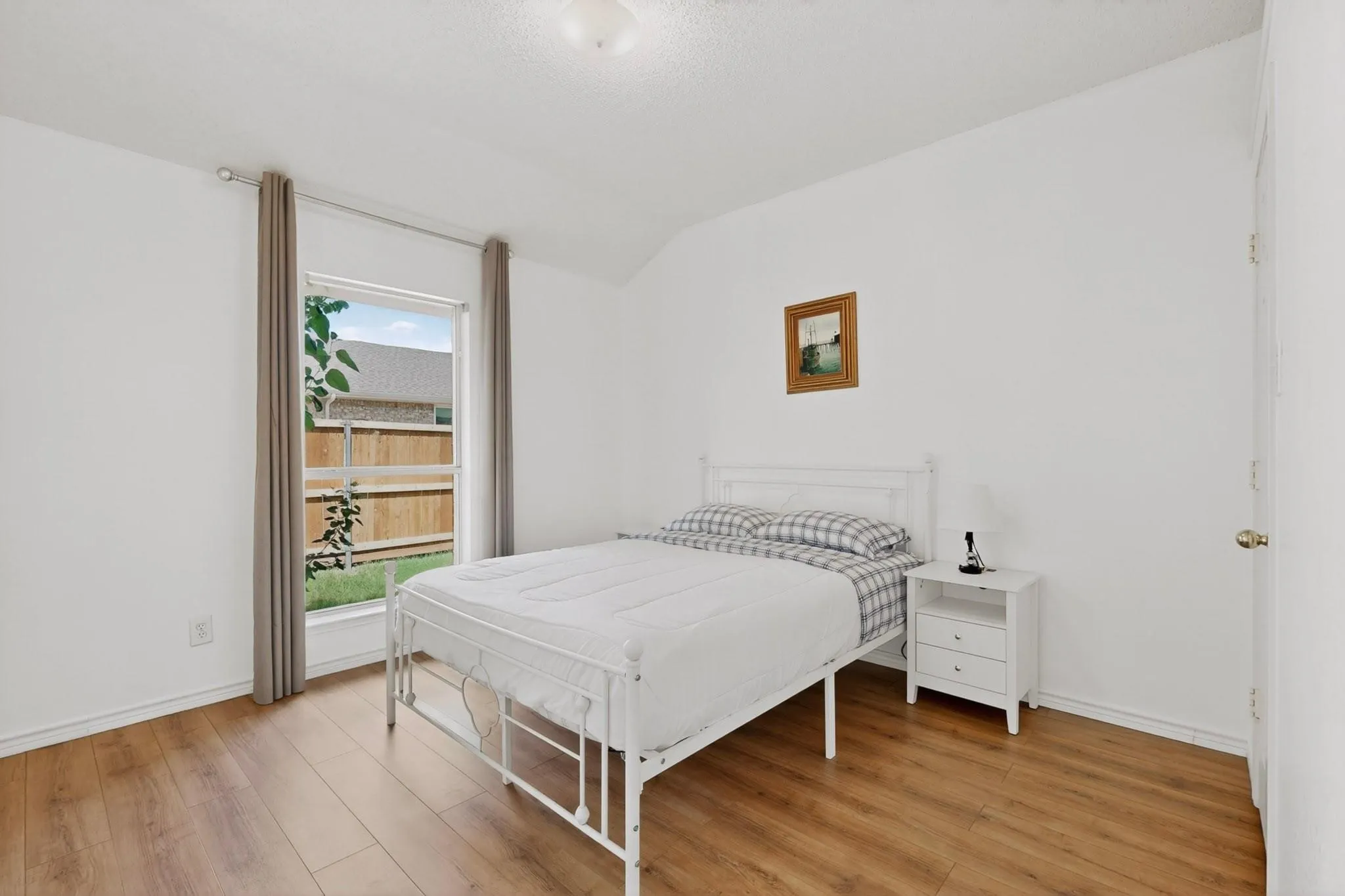 Wood-finish flooring, white painted walls, and a single window with curtain rods