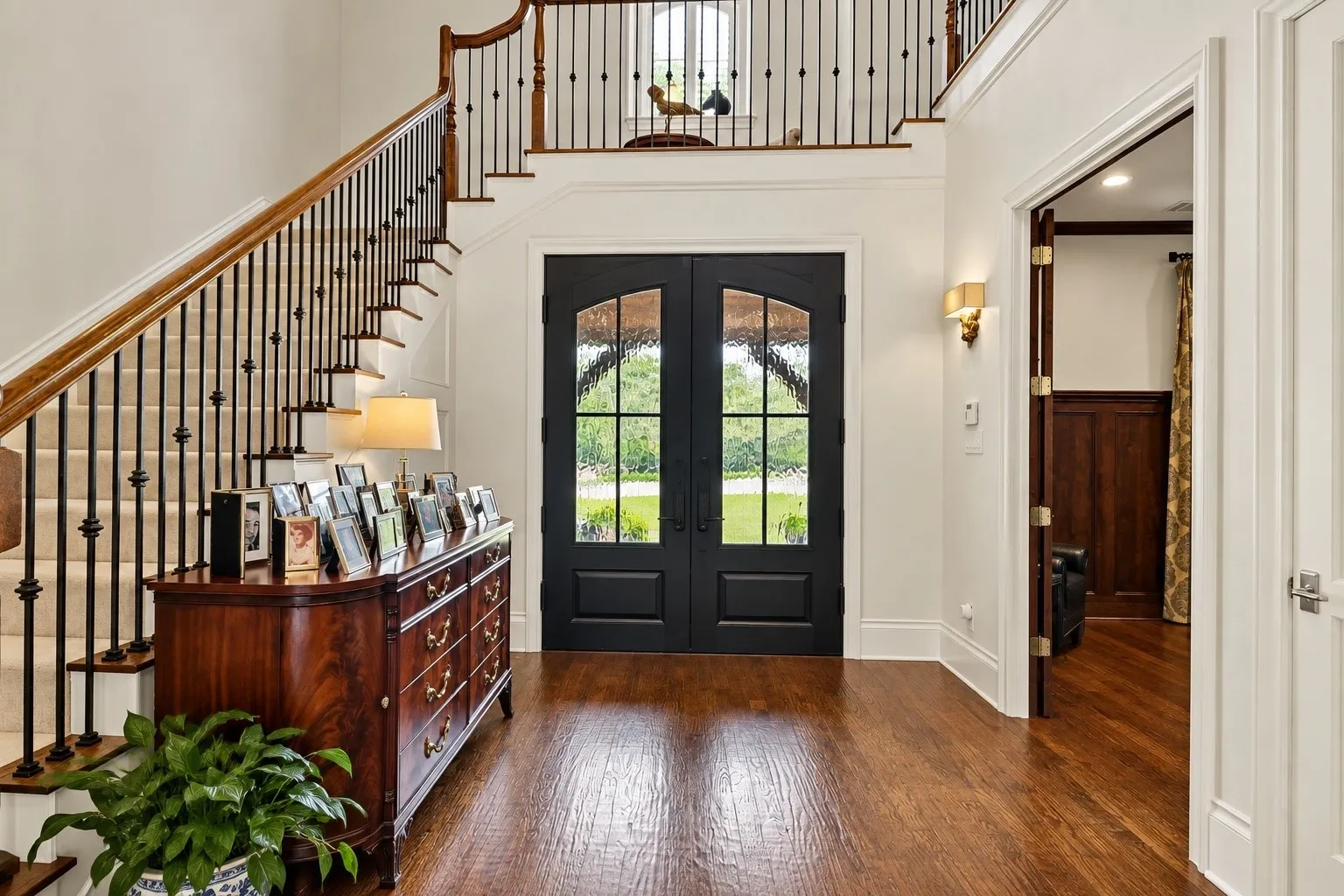 Grand entryway with wood-finish flooring, a carpeted staircase featuring wrought iron and wood railings, and double dark-paneled doors with glass inserts