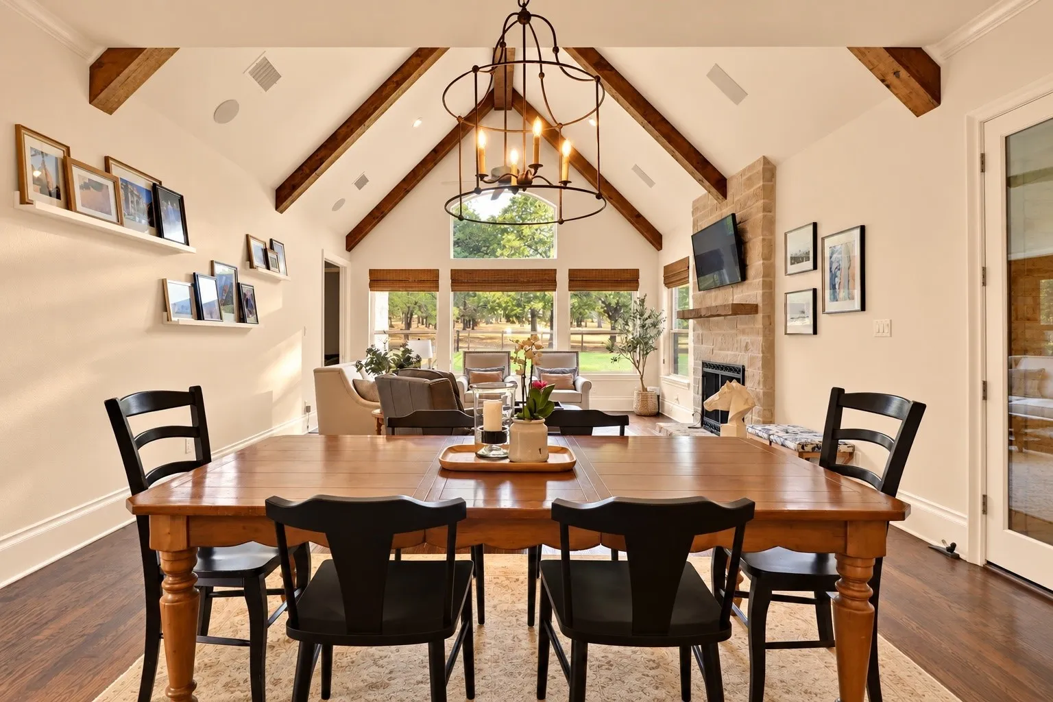 Vaulted ceiling with exposed wood beams, large windows with natural light, stone fireplace, wood-finish flooring, and a glass-paneled door