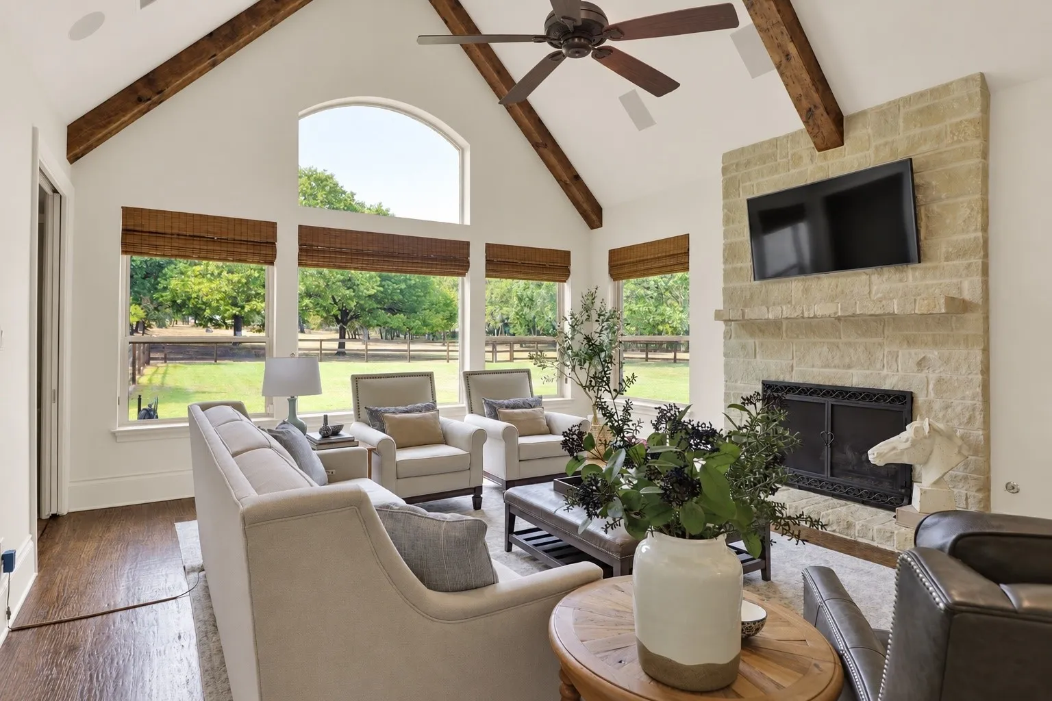 Vaulted ceiling with exposed wood beams, stone-clad fireplace with mantel, large arched window, multiple windows with woven shades, and wood-finish flooring