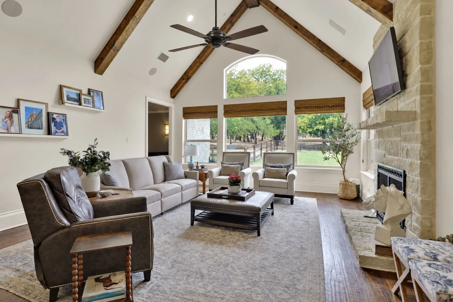 Vaulted ceiling with exposed wood beams, stone-clad fireplace, large windows with arched transom, wood-finish flooring, and recessed lighting