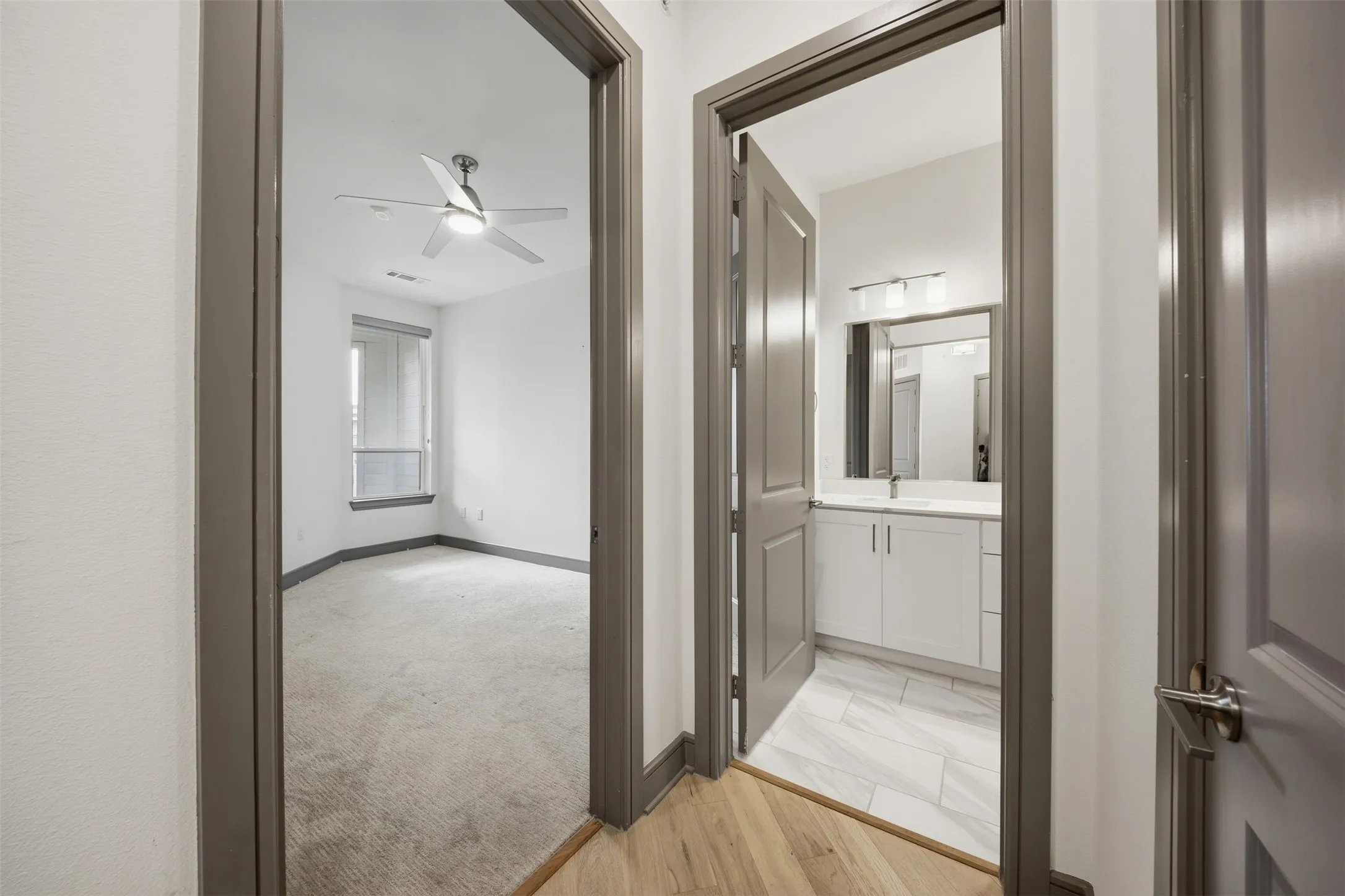 Light wood-finish flooring, light grey carpeting, light grey wall paint, grey interior doors, and modern ceiling fan