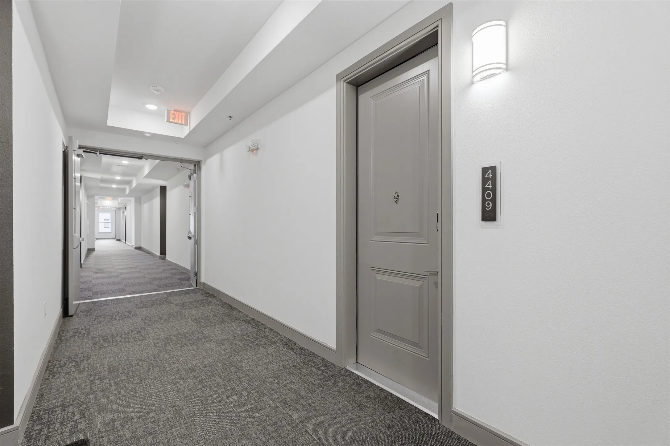 Gray paneled entry door with modern number plate, sconce lighting, and patterned gray carpet