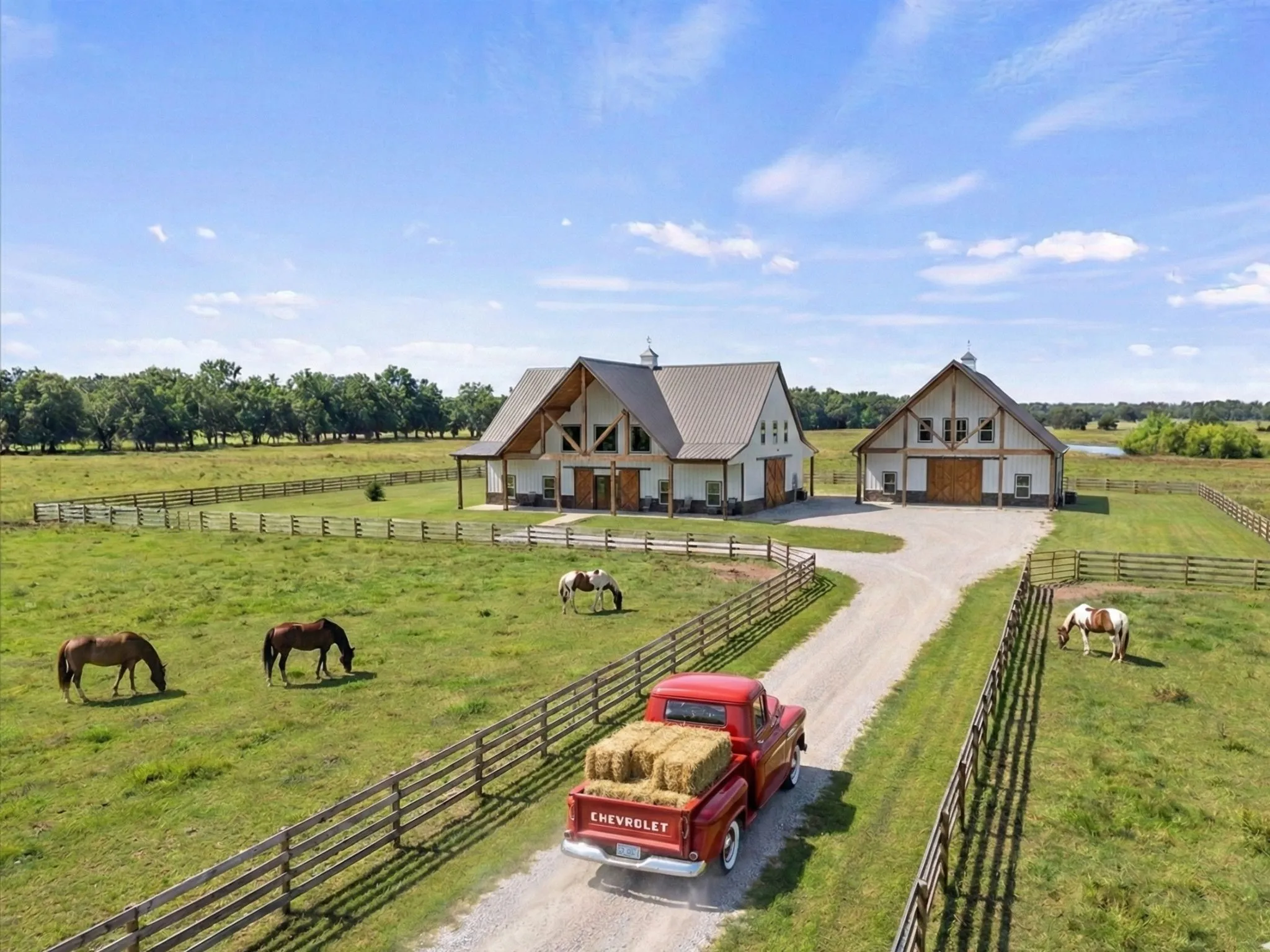Expansive green pastures with wood fencing, a graveled drive, and two gabled barns featuring white siding and wood accents