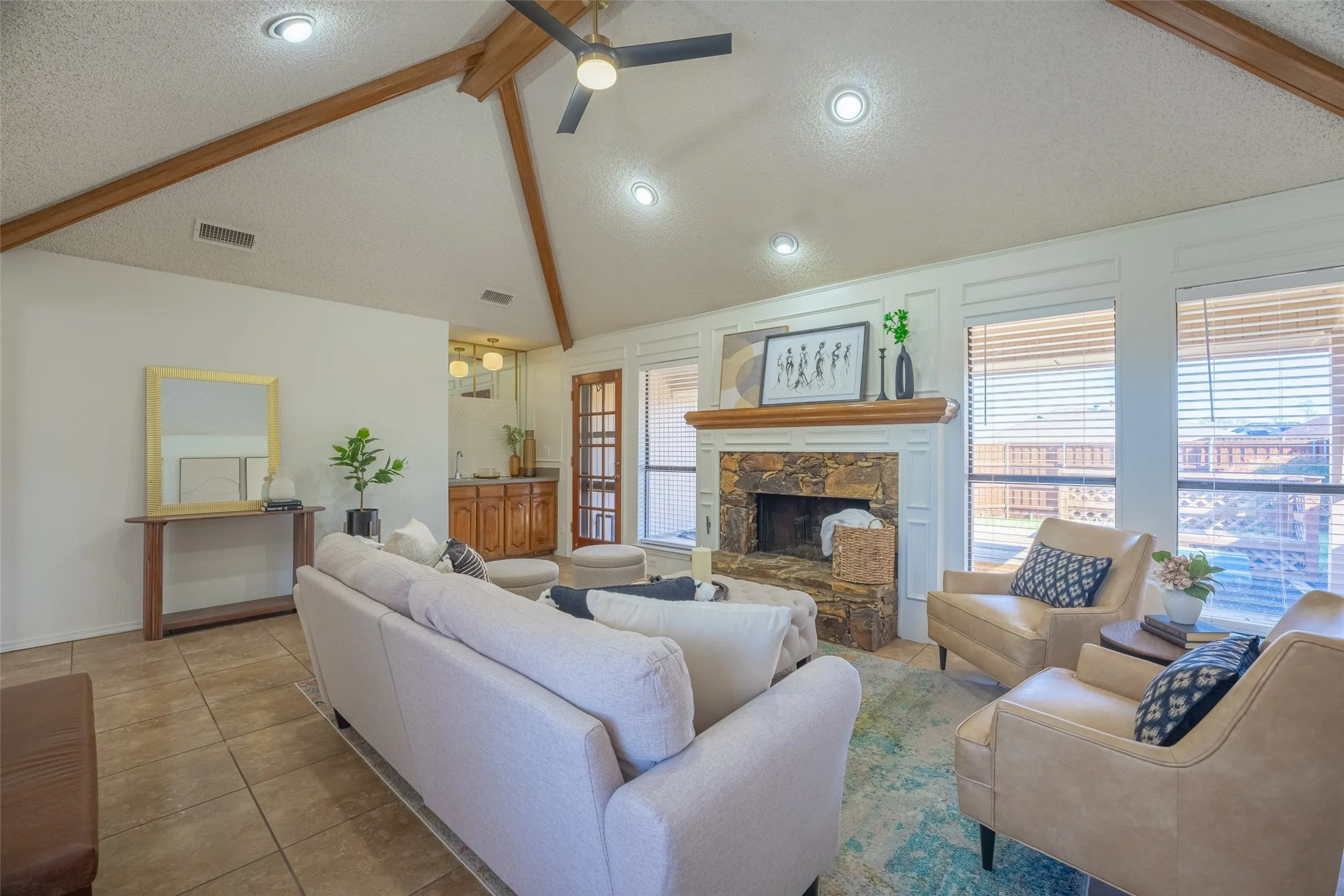 Vaulted ceiling with exposed wood beams and recessed lighting, stone-clad fireplace with white mantel, tile flooring, built-in wet bar with wood cabinetry, and multiple windows with blinds