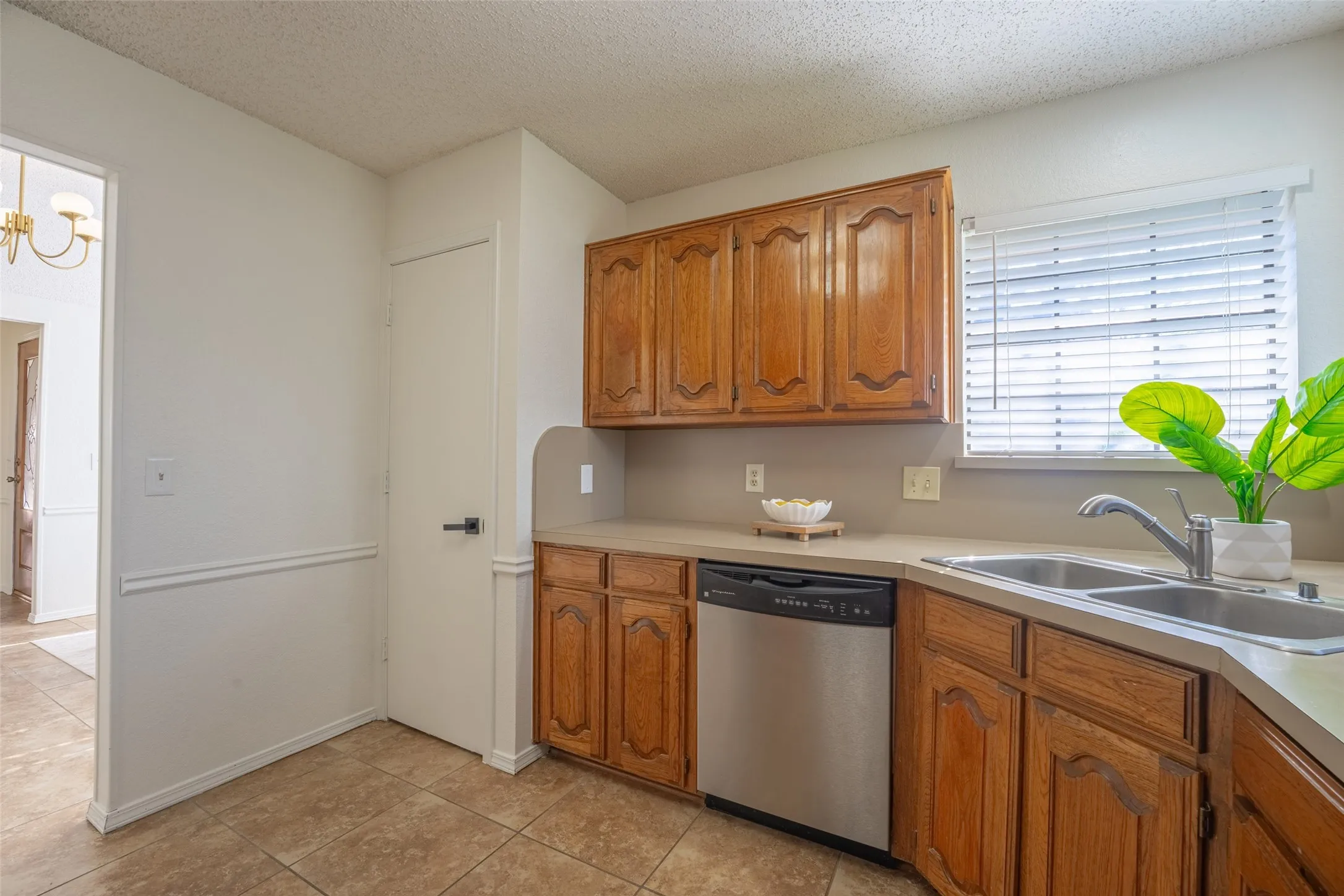 Kitchen featuring wood cabinetry, a stainless steel dishwasher, a double basin sink with a chrome faucet, light-toned countertops, and tiled flooring
