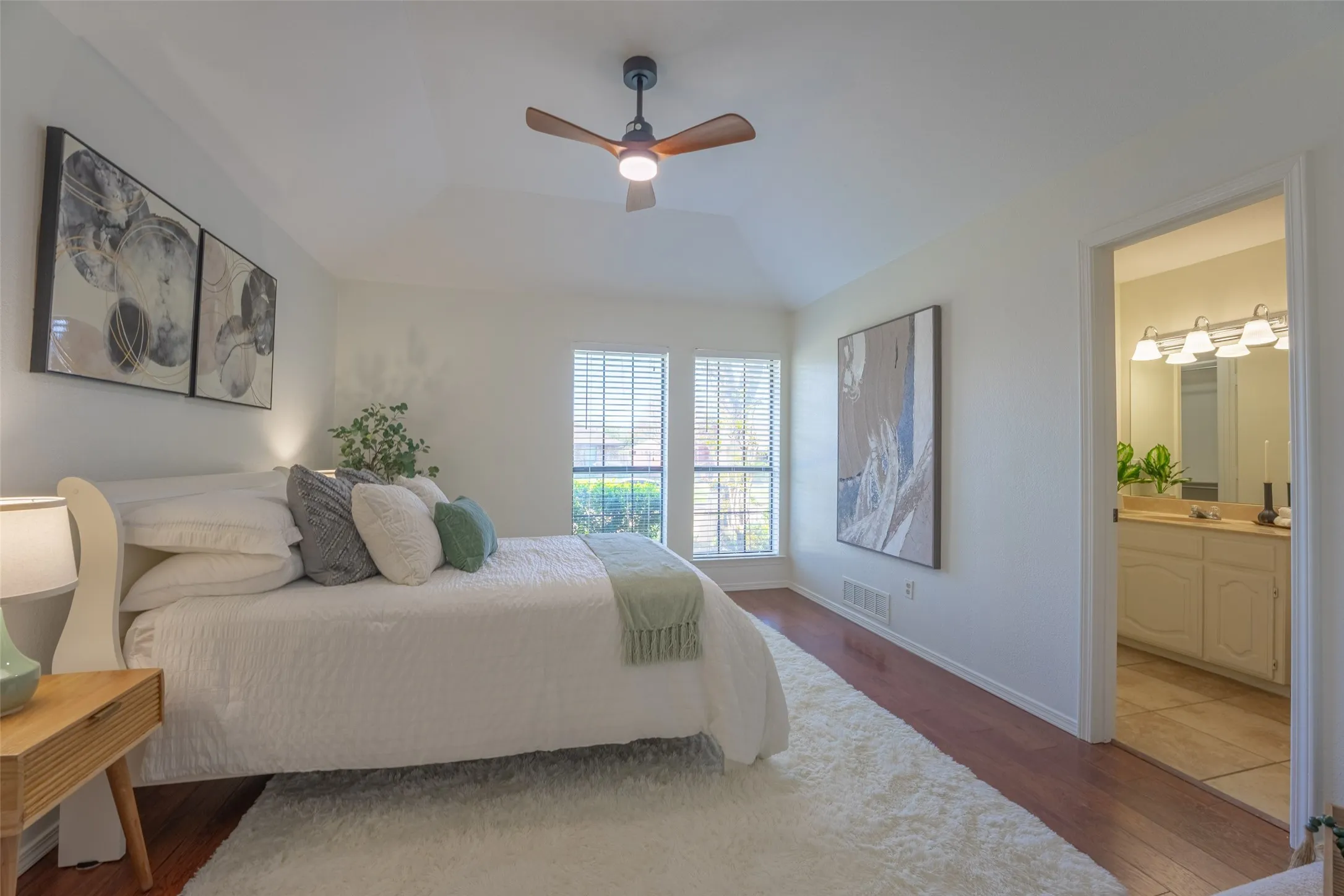 Vaulted ceiling with a ceiling fan, dual windows with blinds, wood-finish flooring, and an adjoining bathroom with a vanity and mirror