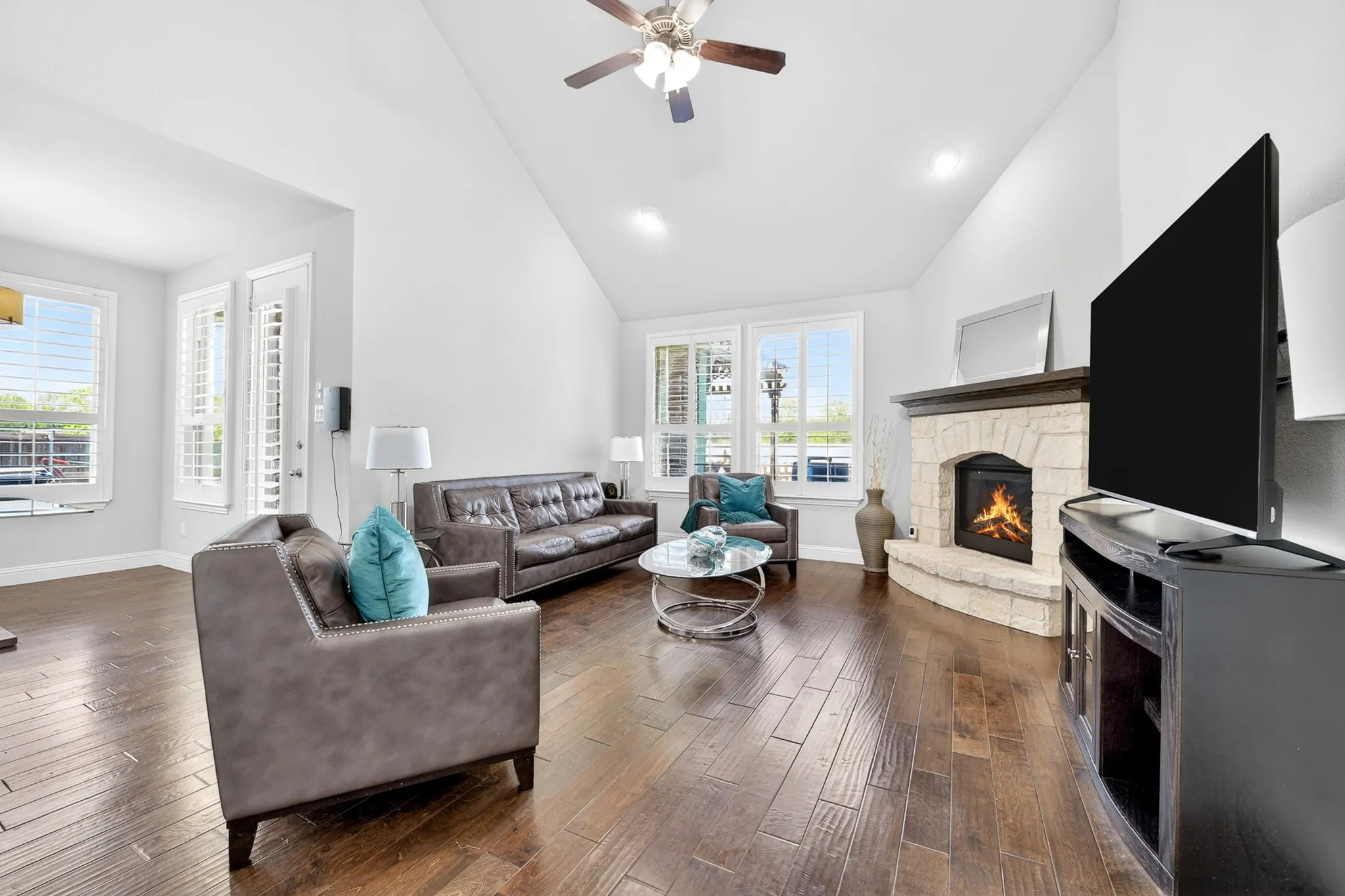 Vaulted ceilings, wood-finish flooring, stone fireplace with hearth, white window shutters, and a ceiling fan with integrated lighting