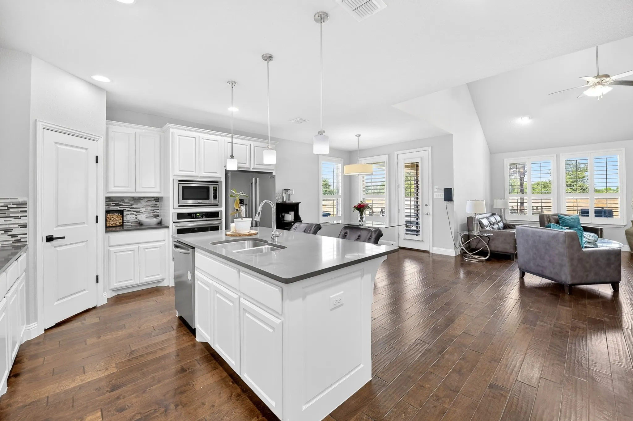 Wood-finish flooring throughout, kitchen with white cabinetry and stainless steel appliances, central island with sink, living area with vaulted ceiling, and multiple windows with plantation shutters