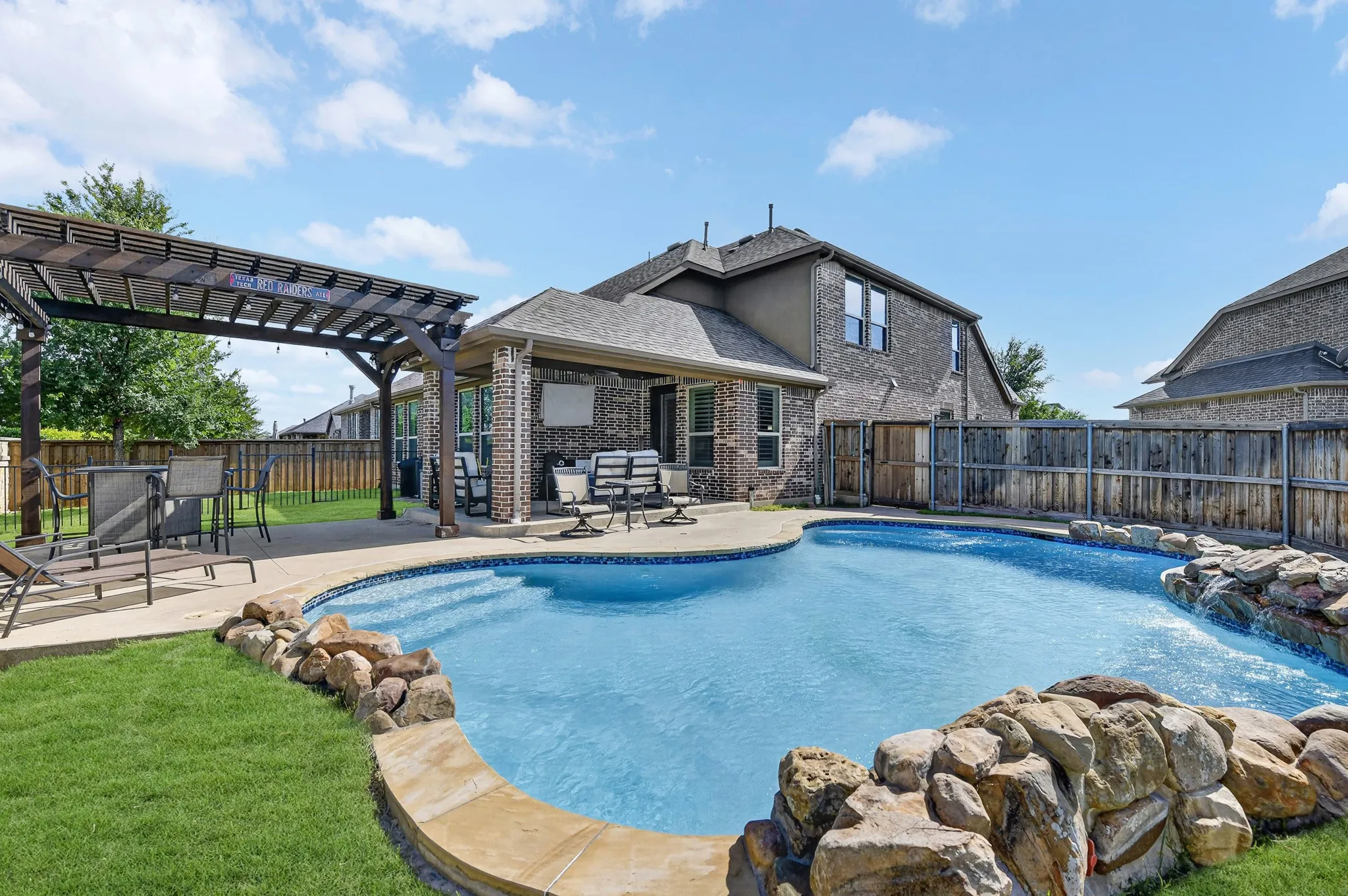 Curved swimming pool with rock waterfall feature, adjacent to a covered patio and a separate pergola with concrete decking