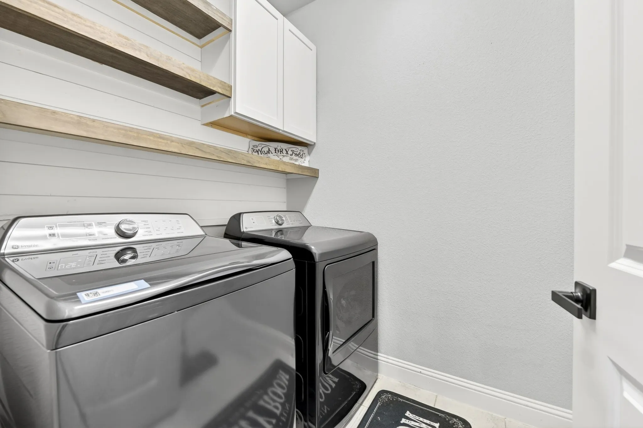 Laundry area featuring shiplap wall treatment, two wood-finish floating shelves, and white upper cabinetry