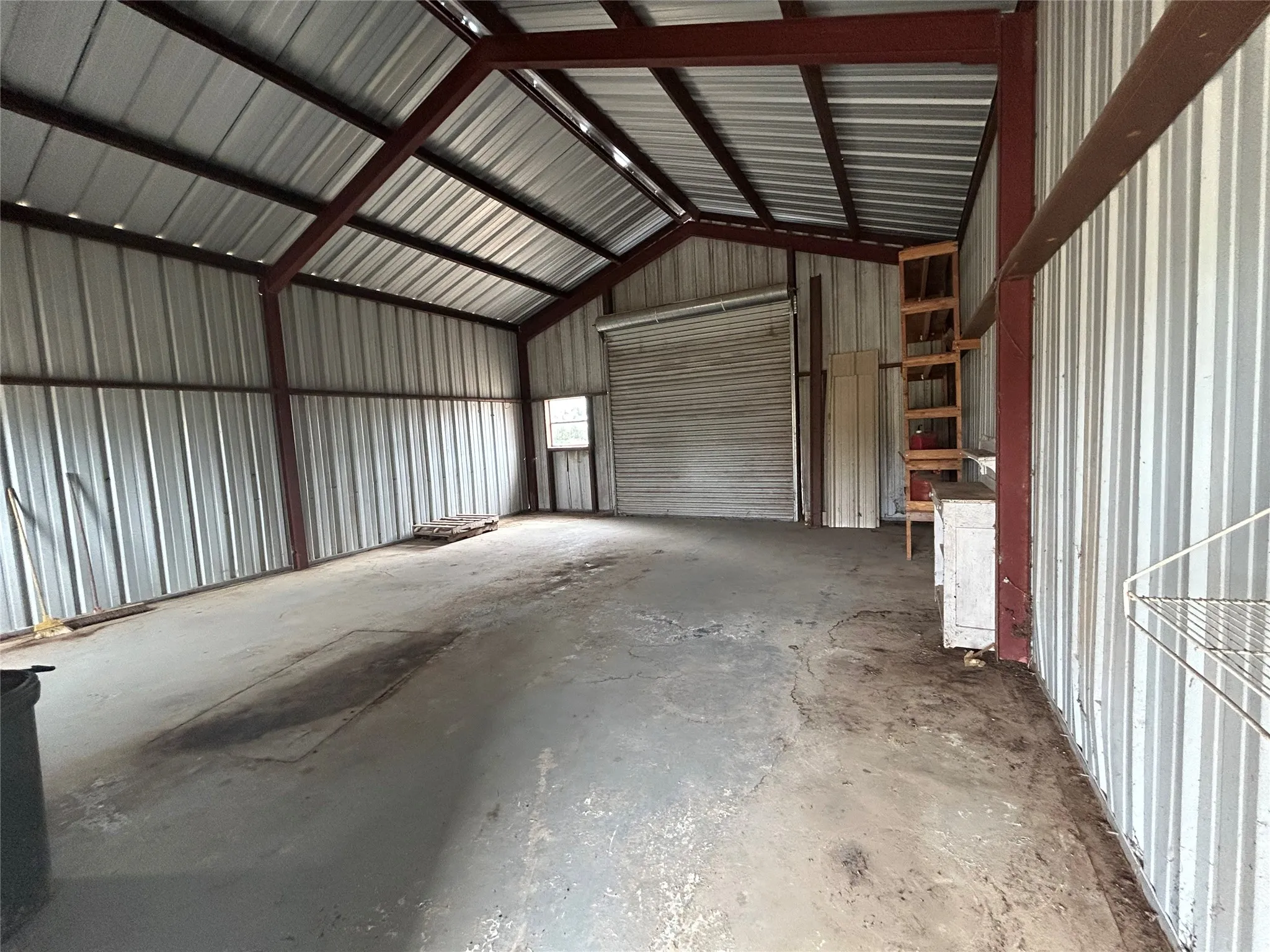 Expansive metal building featuring a vaulted ceiling with exposed structural beams, corrugated metal walls, a large roll-up door, a service door, and a built-in shelving unit