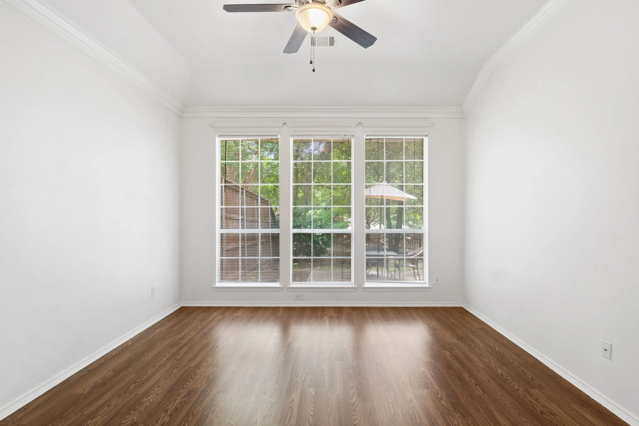 Vaulted ceiling with crown molding, ceiling fan, wood-finish flooring, and a bank of three large windows