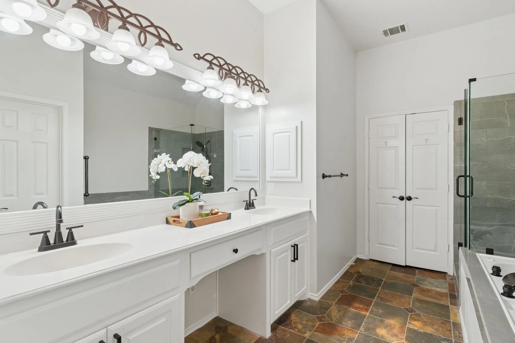 Dual vanity with integrated sinks and black-finish faucets, expansive framed mirror, tiled flooring, and a glass-enclosed shower with dark tile surround
