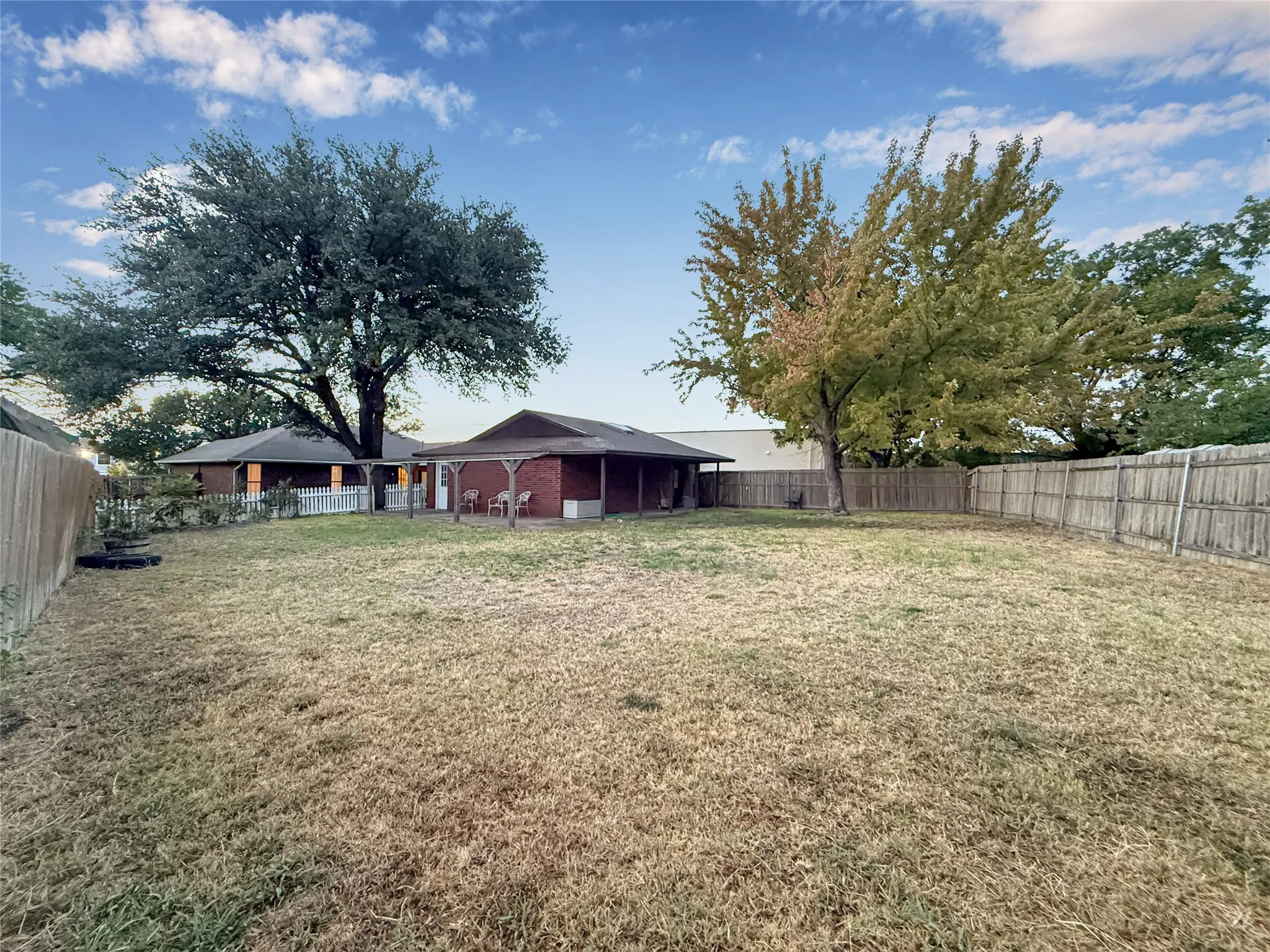 Expansive backyard featuring a privacy fence, mature trees, and a covered patio area with a brick exterior