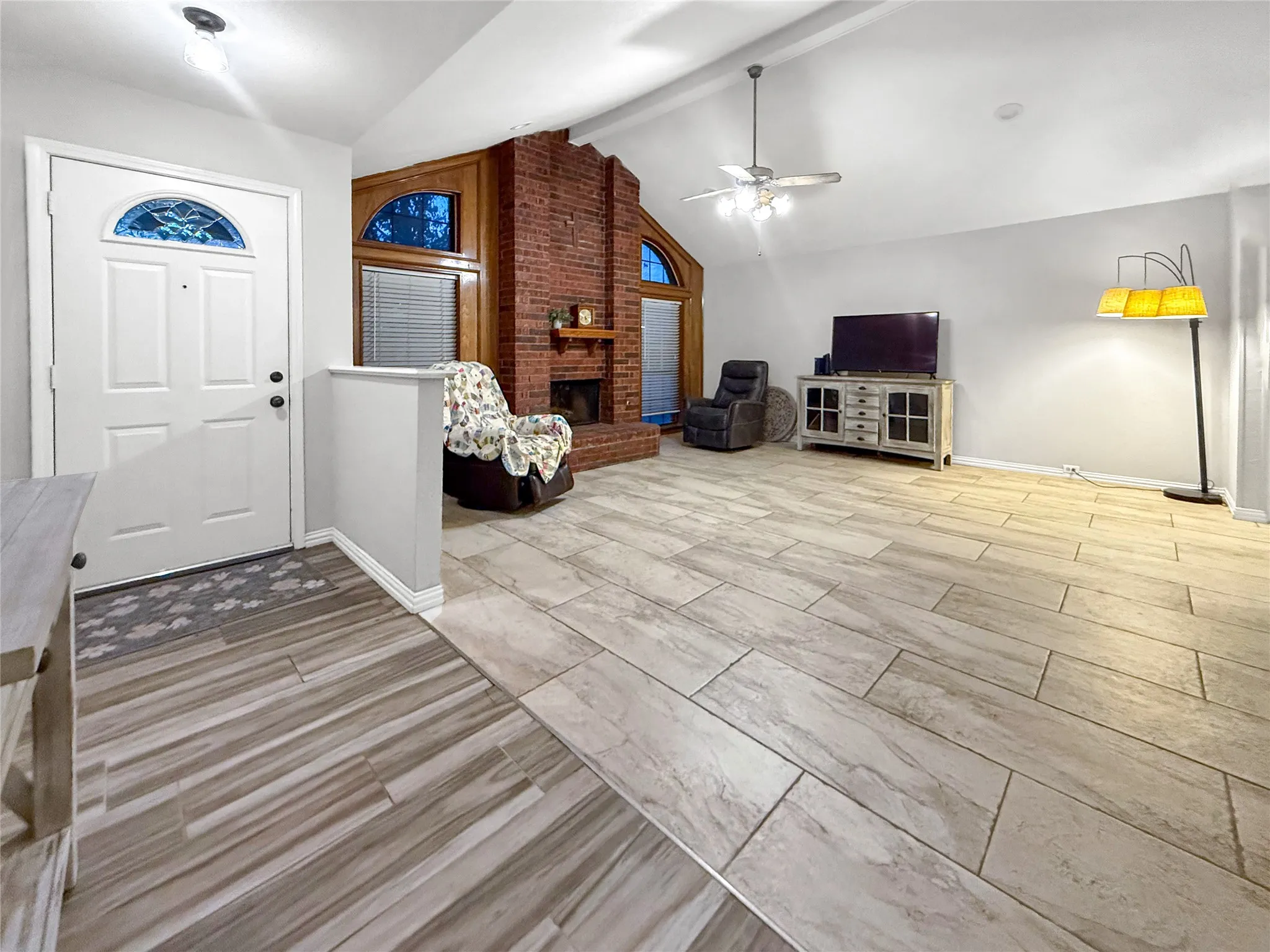 Vaulted ceilings with a ceiling fan, brick fireplace with wood mantel, arched windows with wood trim, and tile flooring