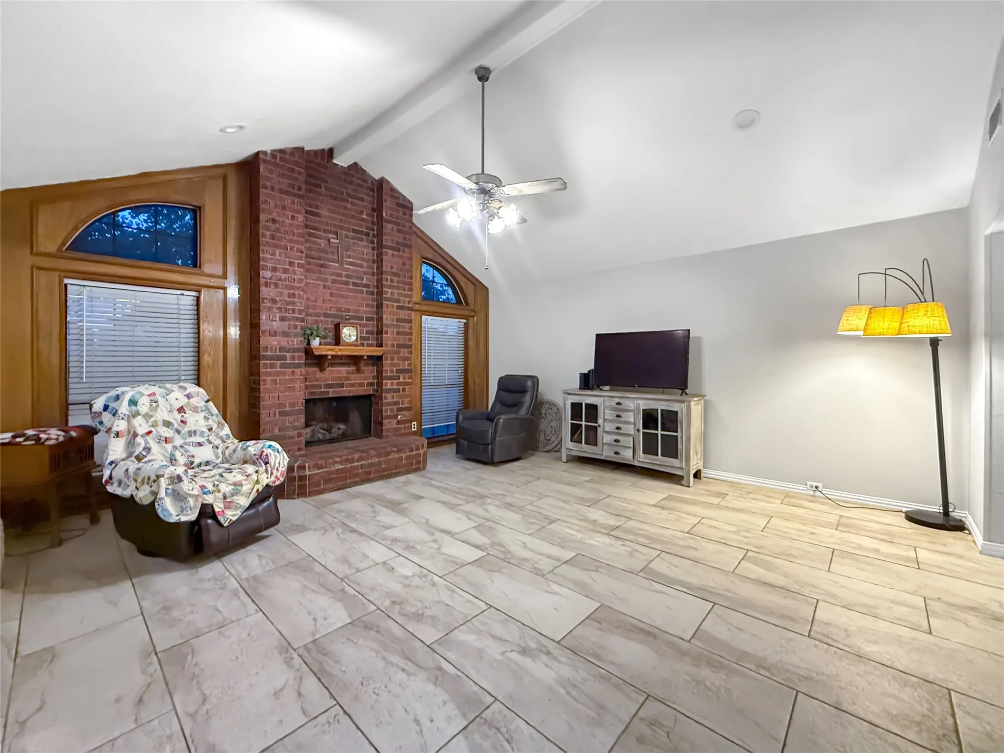 Vaulted ceiling with a ceiling fan, brick fireplace with a wood mantle, wood-paneled accents, and tile flooring