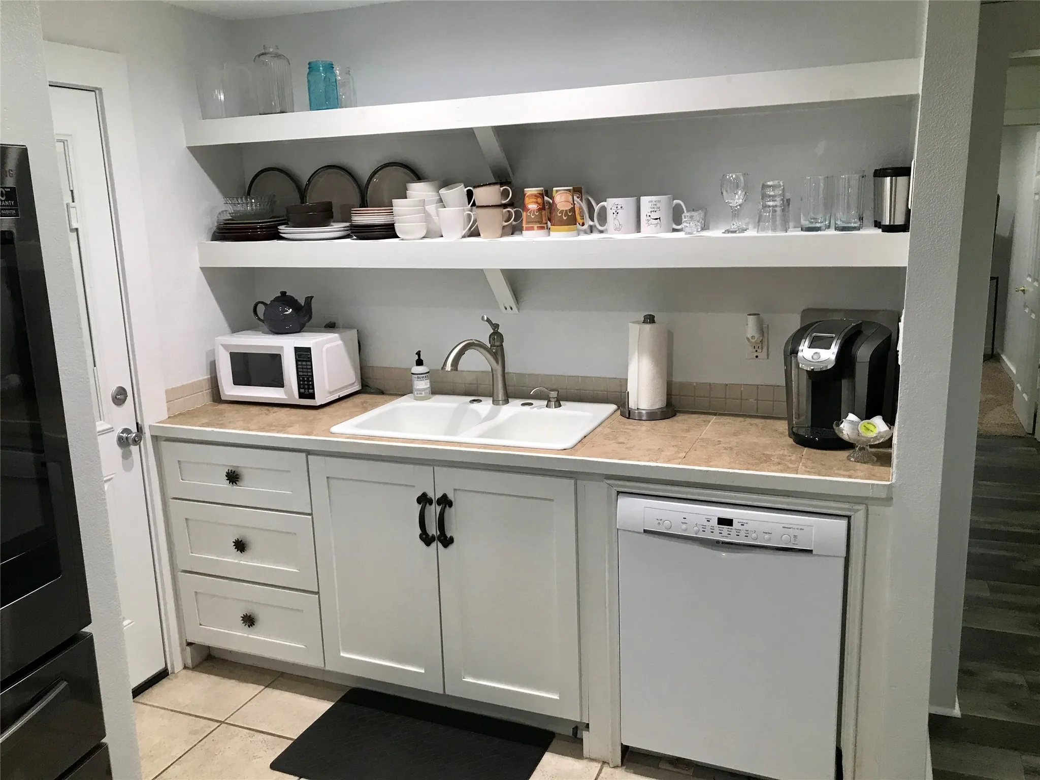 White cabinetry with black hardware, tile flooring, built-in dishwasher, dual basin sink, and open shelving