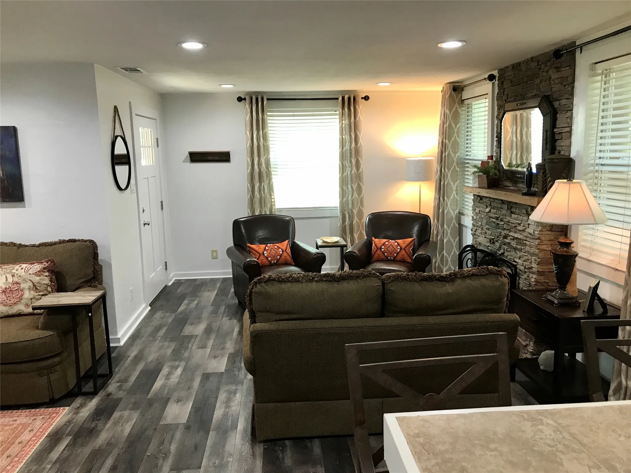 Wood-finish flooring, recessed ceiling lighting, white interior door, stone-clad fireplace, and multiple windows with blinds