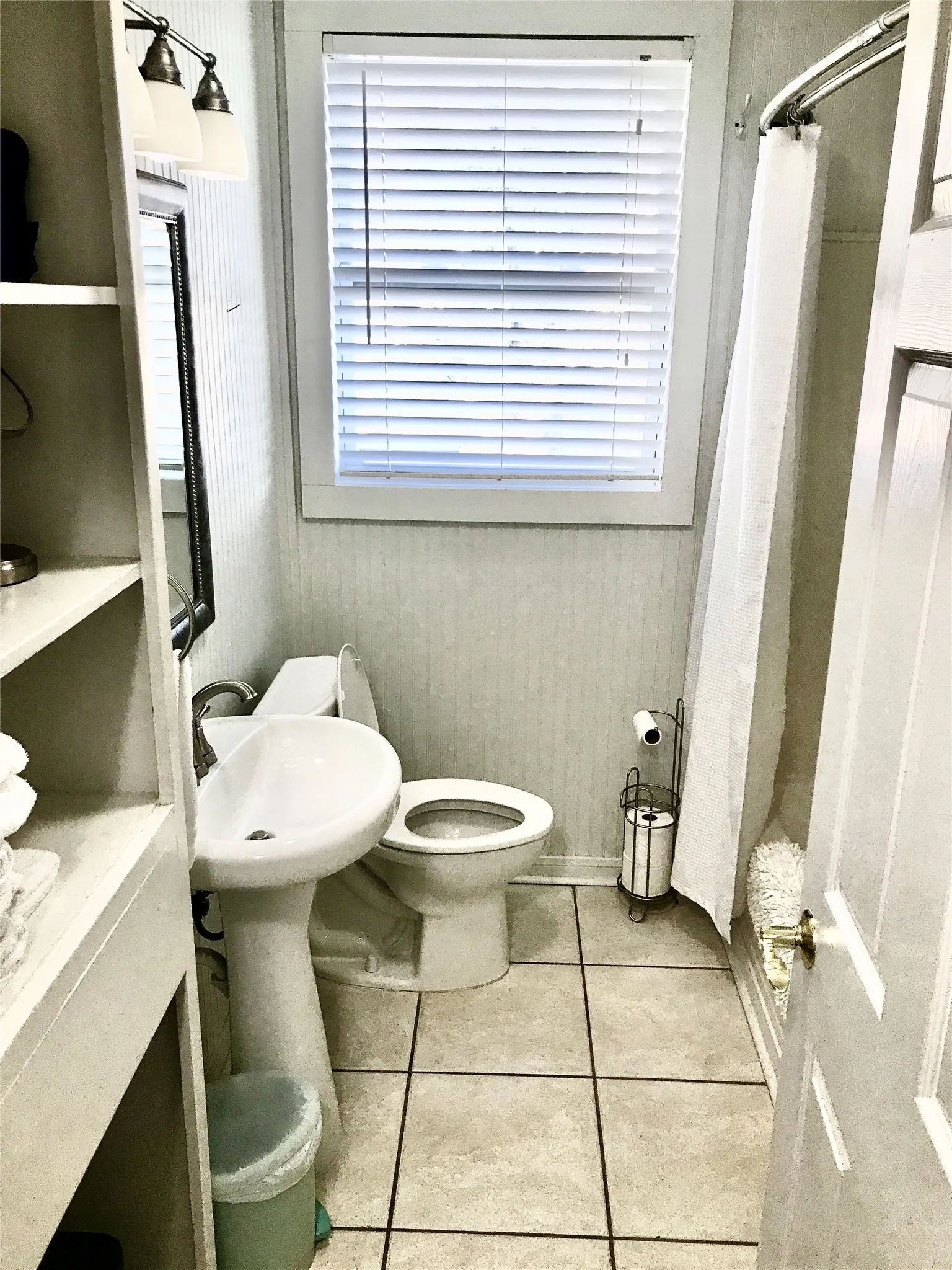 Pedestal sink with chrome faucet, ceramic tile flooring, built-in shelving, white window blinds, and a shower/tub combination
