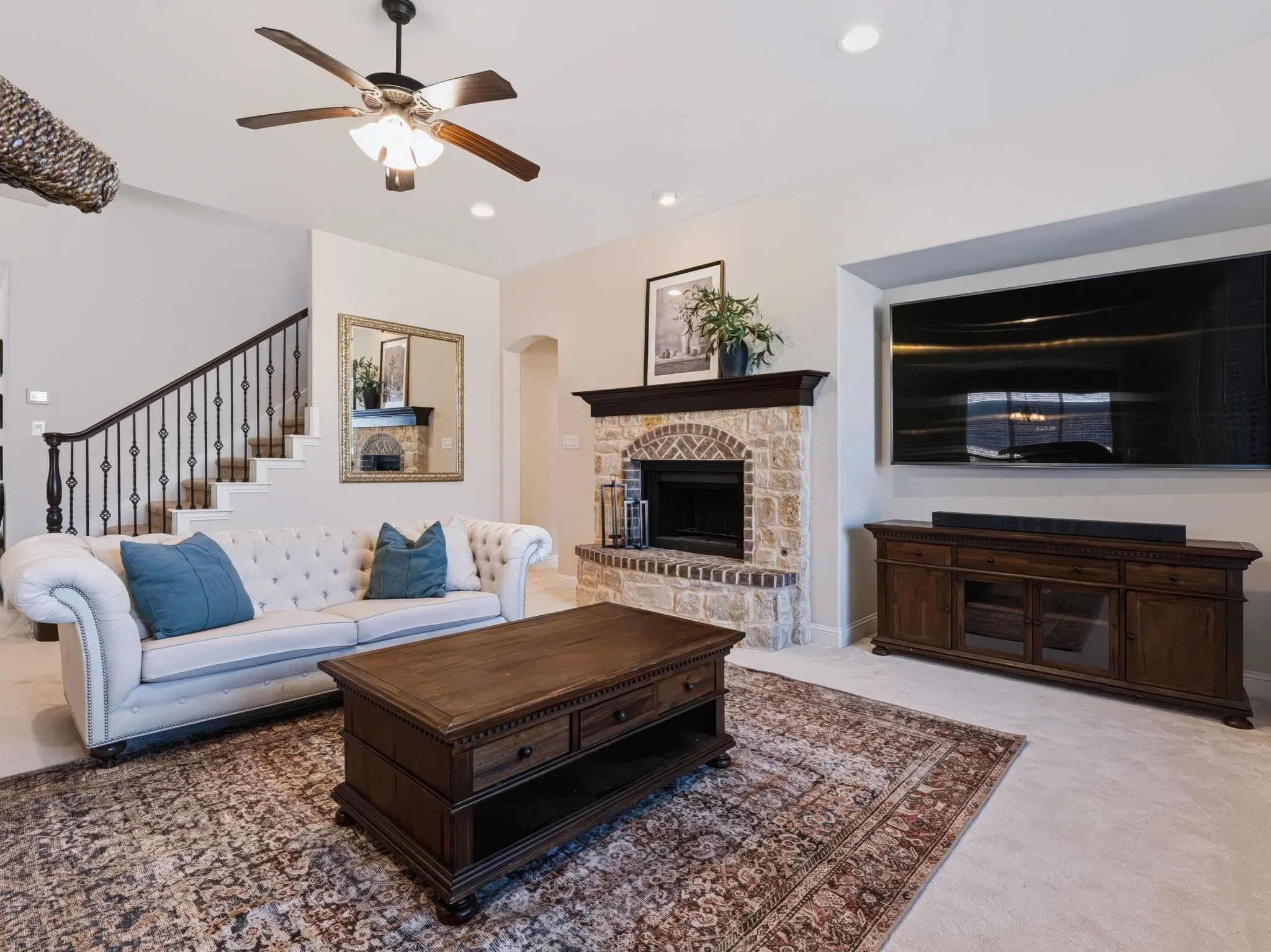 Stone fireplace with dark wood mantel, recessed lighting, ceiling fan, and staircase with wrought iron balusters