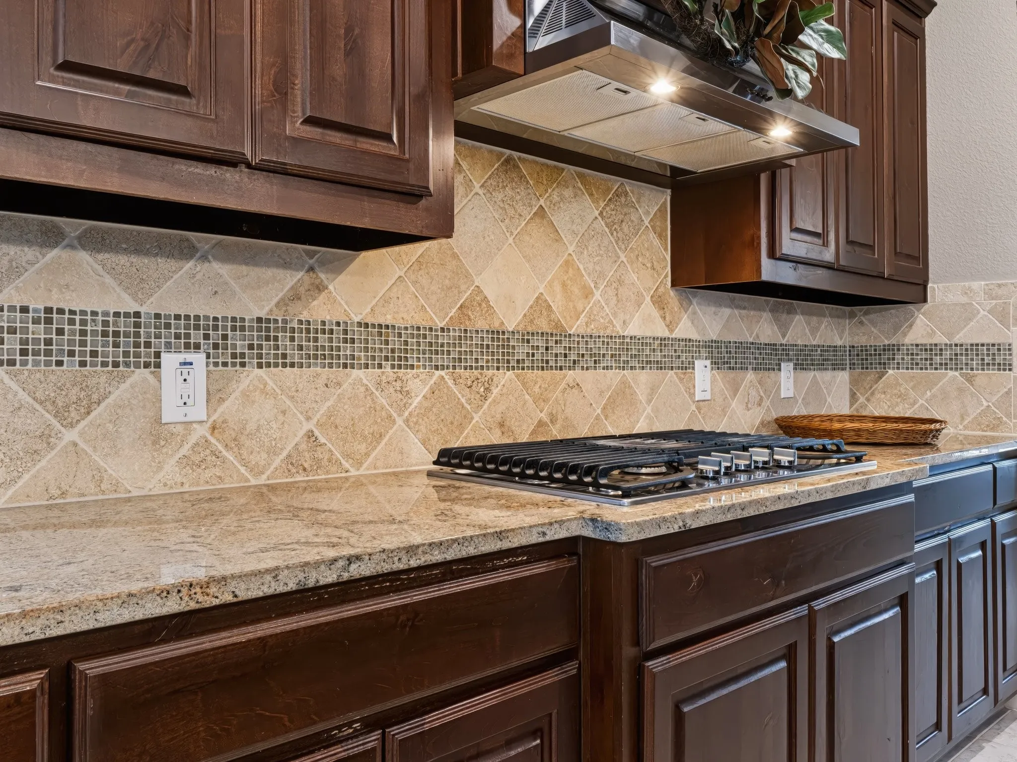 Integrated gas cooktop with a range hood, complemented by a tiled backsplash featuring a mosaic accent band, rich wood-finish cabinetry, and solid surface countertops