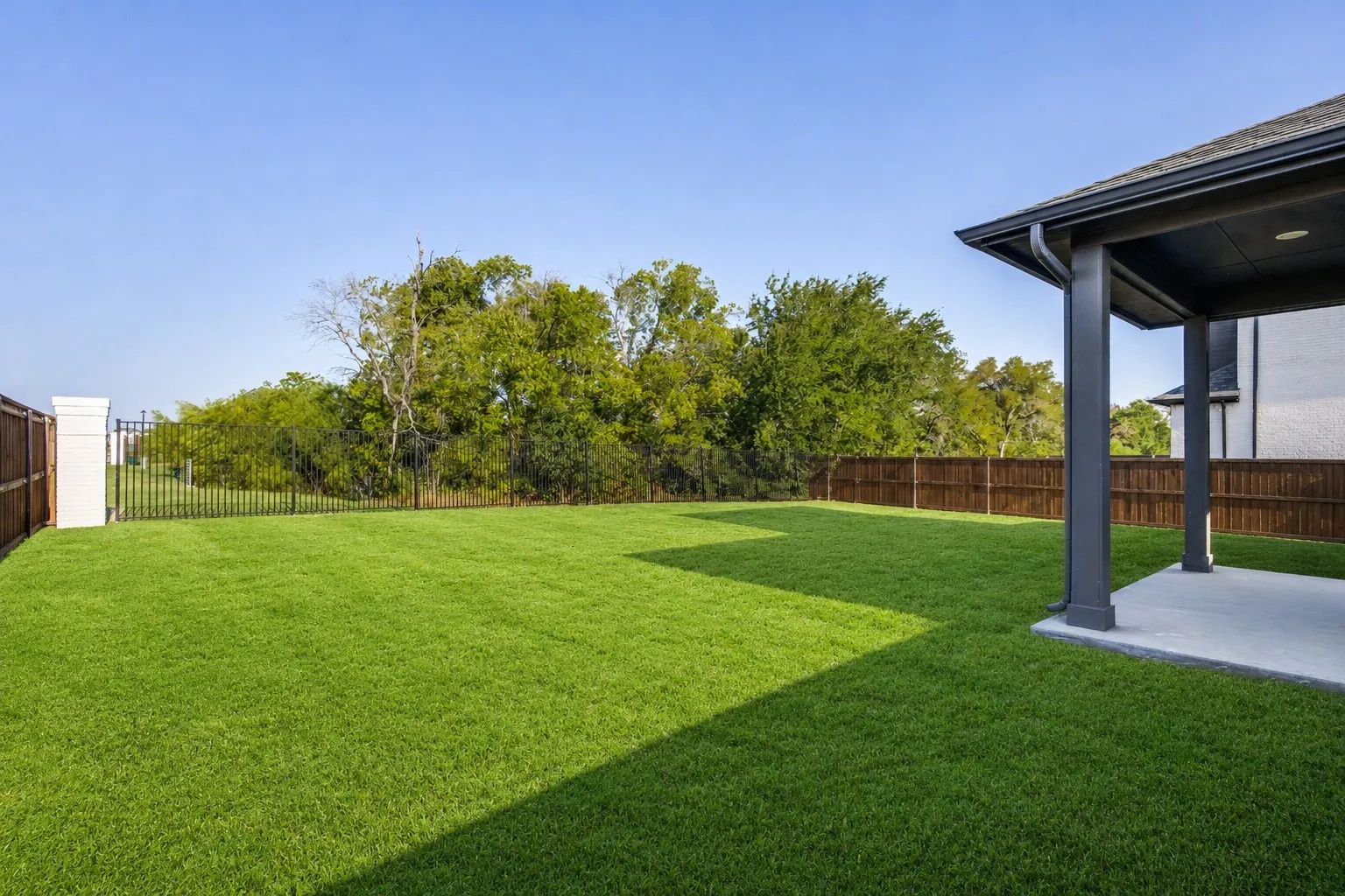 Expansive lawn with a covered concrete patio, wood privacy fencing, and a black wrought iron fence